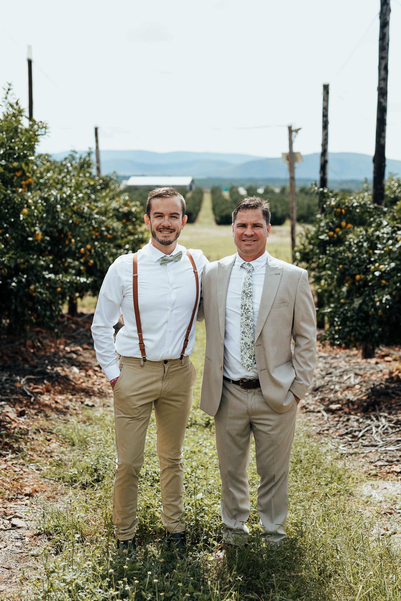 Two people in formal attire stand together on path through citrus grove.