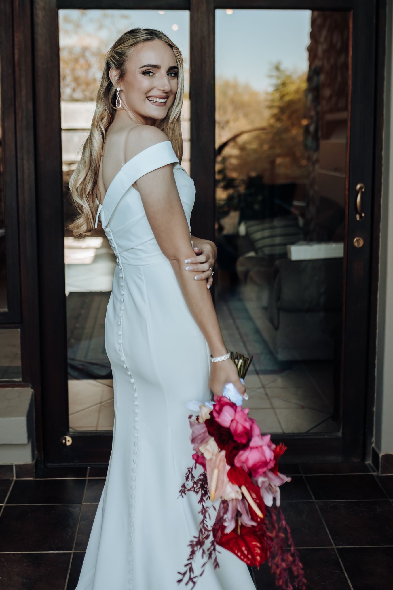 Side profile of bride in fitted white gown holding dramatic red and pink cascading bouquet.