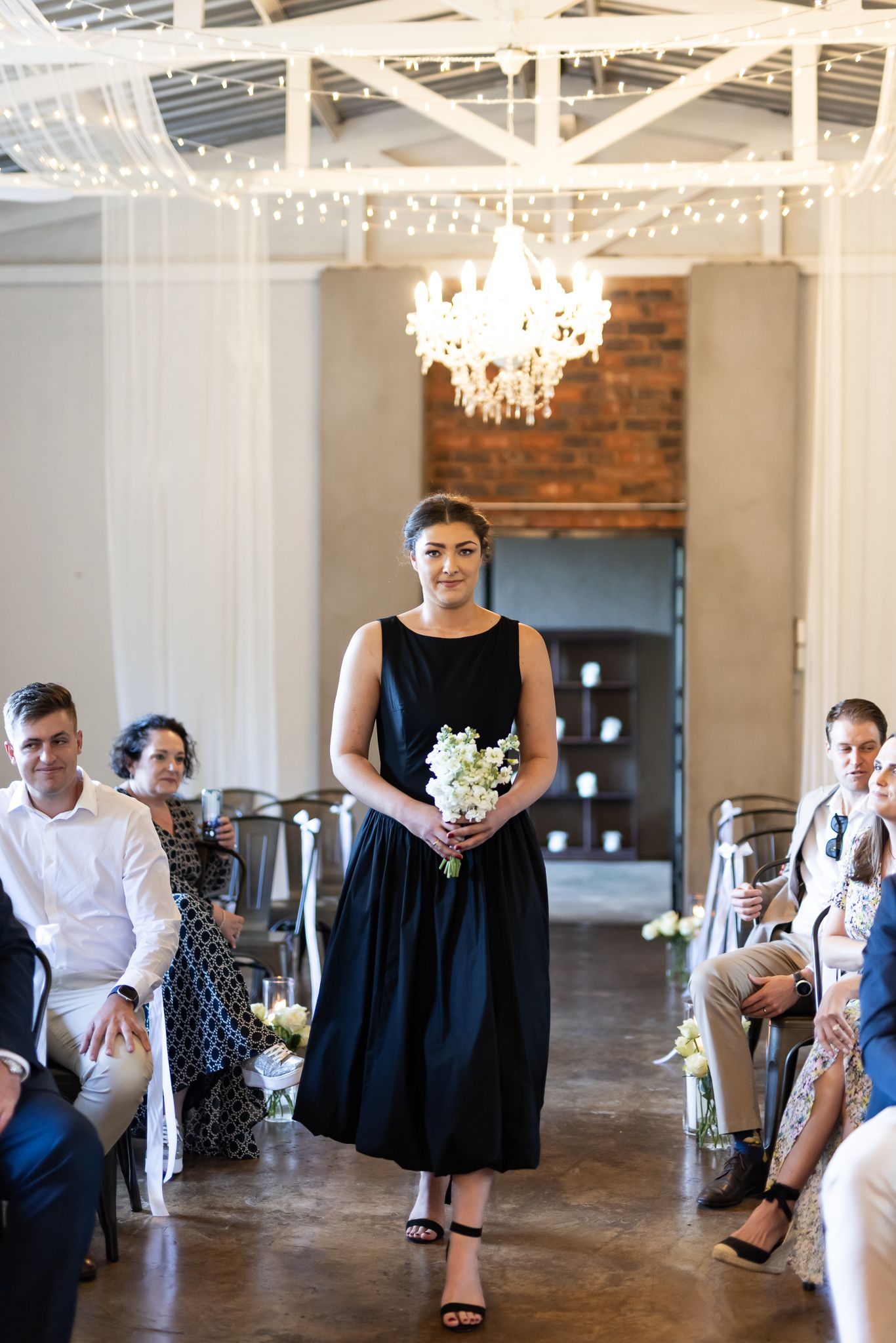 Bridesmaid walks ceremony aisle in black dress under crystal chandelier in industrial venue.