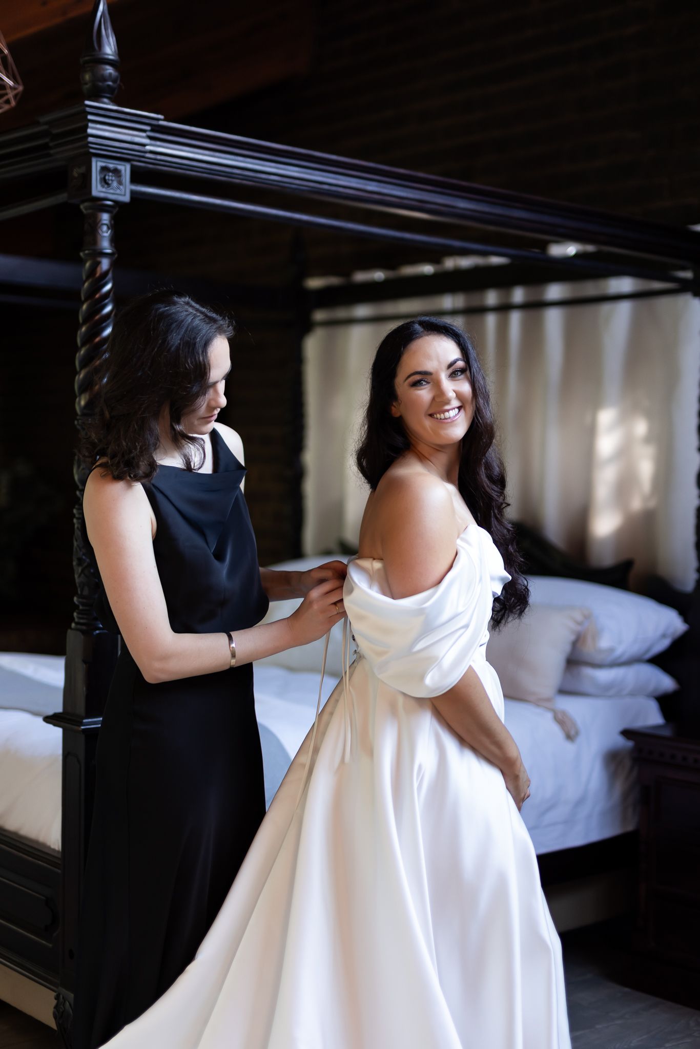 Getting ready scene with white wedding dress being adjusted in bedroom with four poster bed.