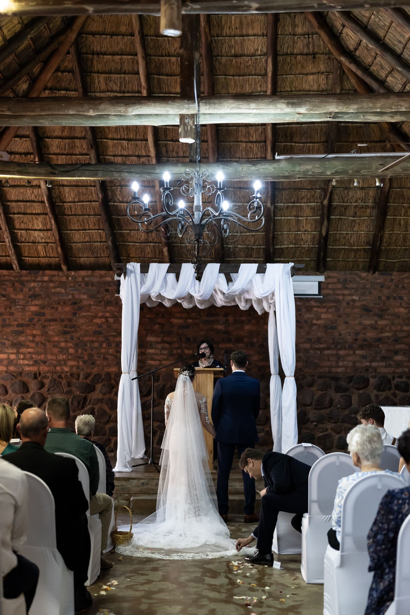 Wedding ceremony setup with white draped arch and chandelier in rustic brick venue with thatched roof.