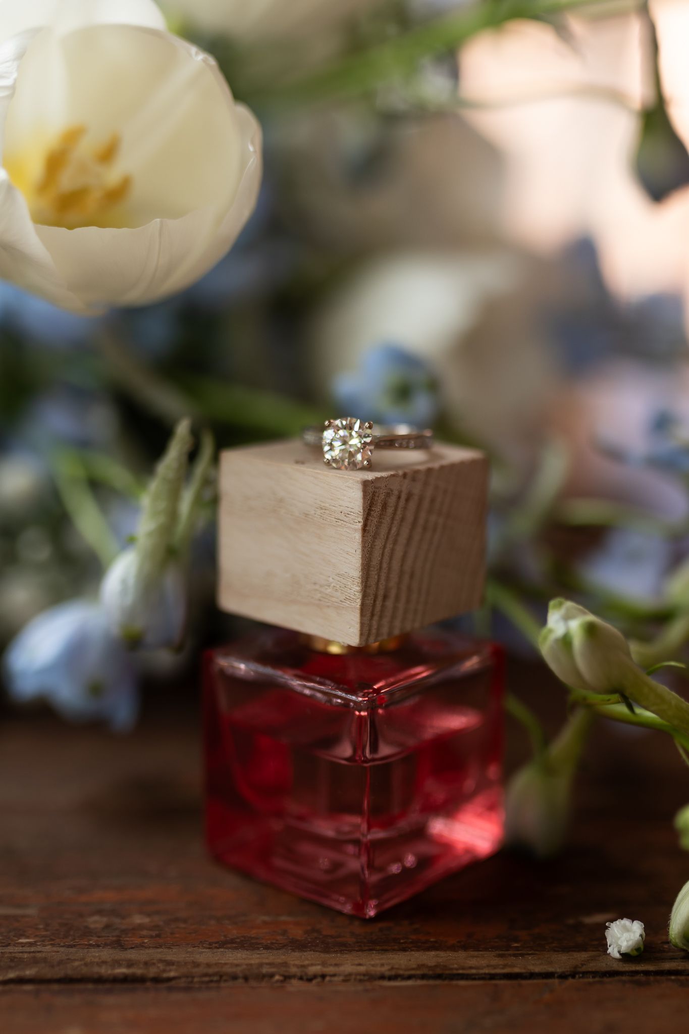 Diamond ring on wooden block next to red perfume bottle and white tulip.
