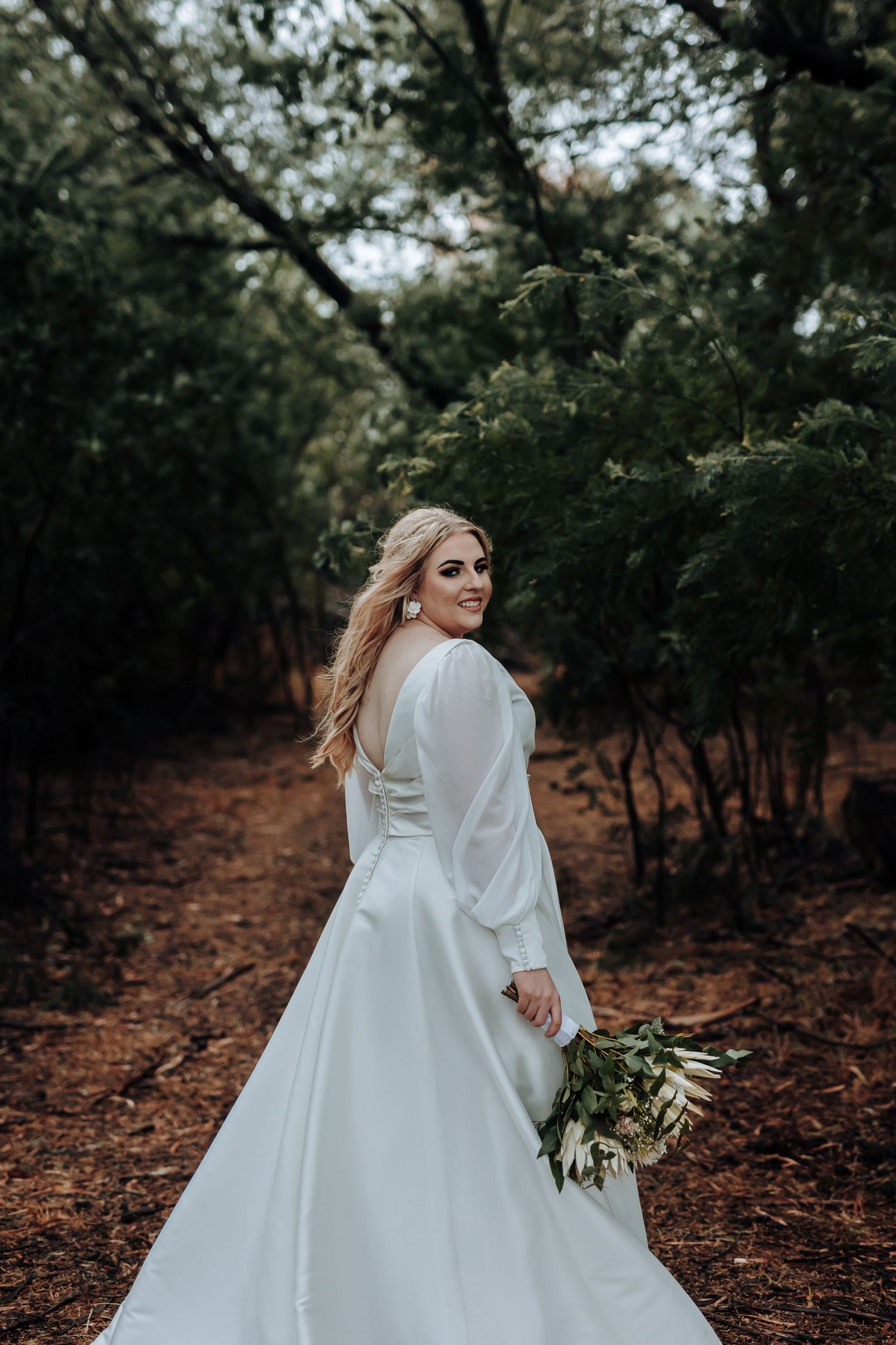 Bride in elegant long-sleeved white dress poses with bouquet on forest path.