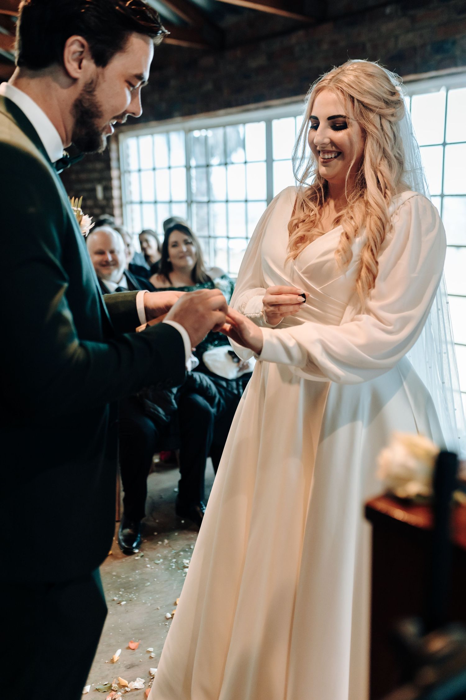 Intimate wedding ceremony moment with couple exchanging rings in front of exposed brick and windows.