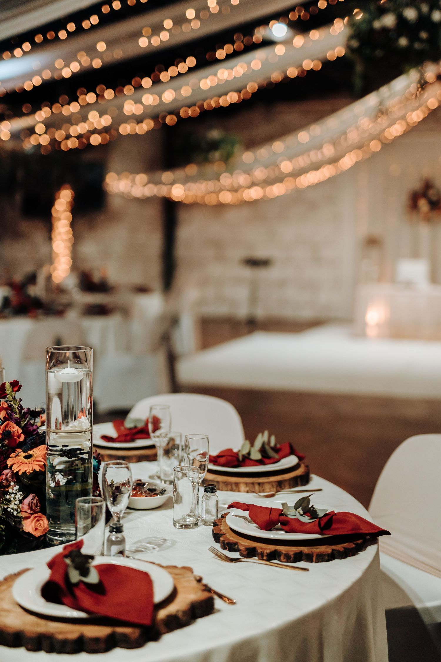 Wedding reception table with white linens, burgundy accents, and rustic wooden charger plates under string lights.