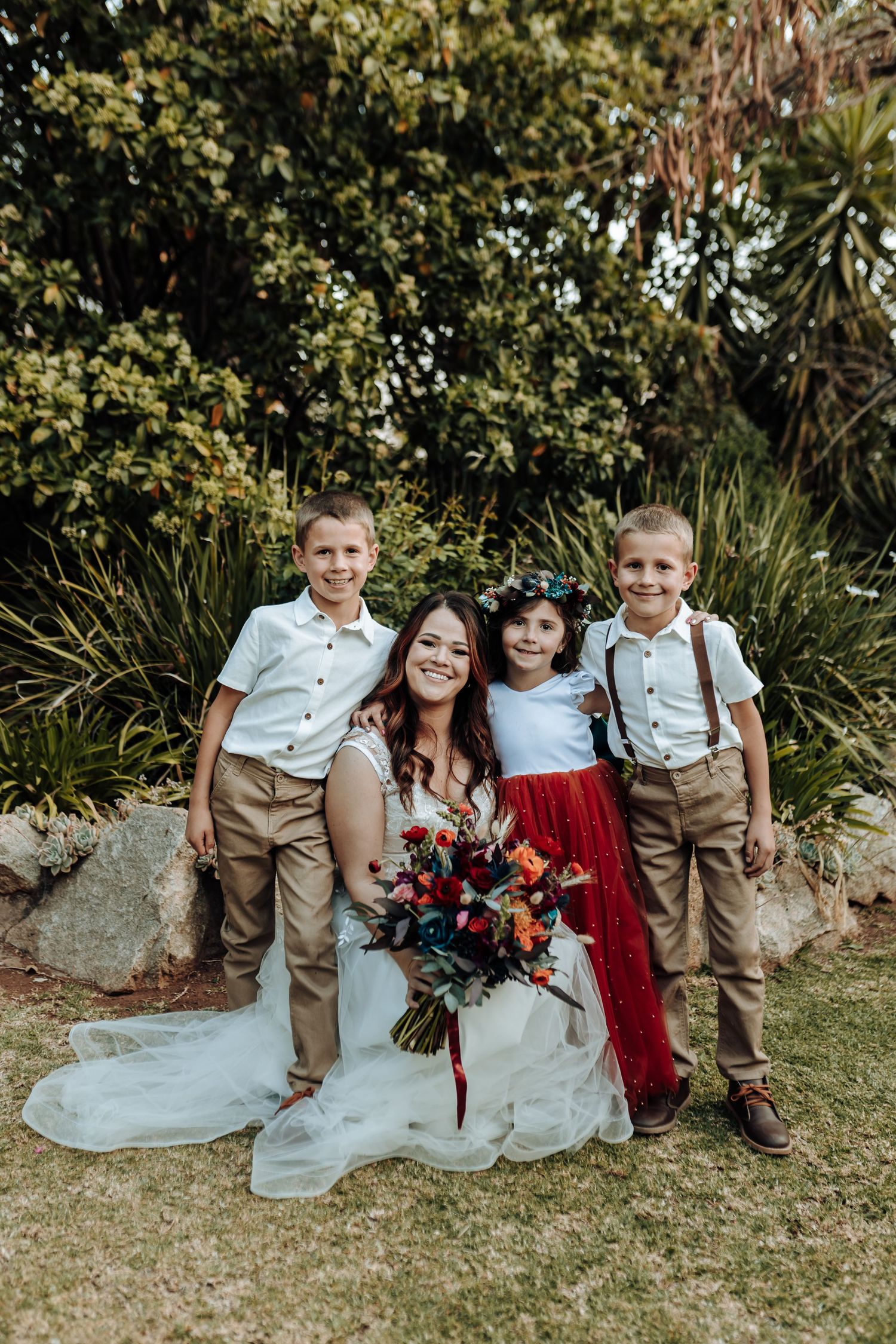 Bride poses with children in coordinated wedding attire against garden backdrop.