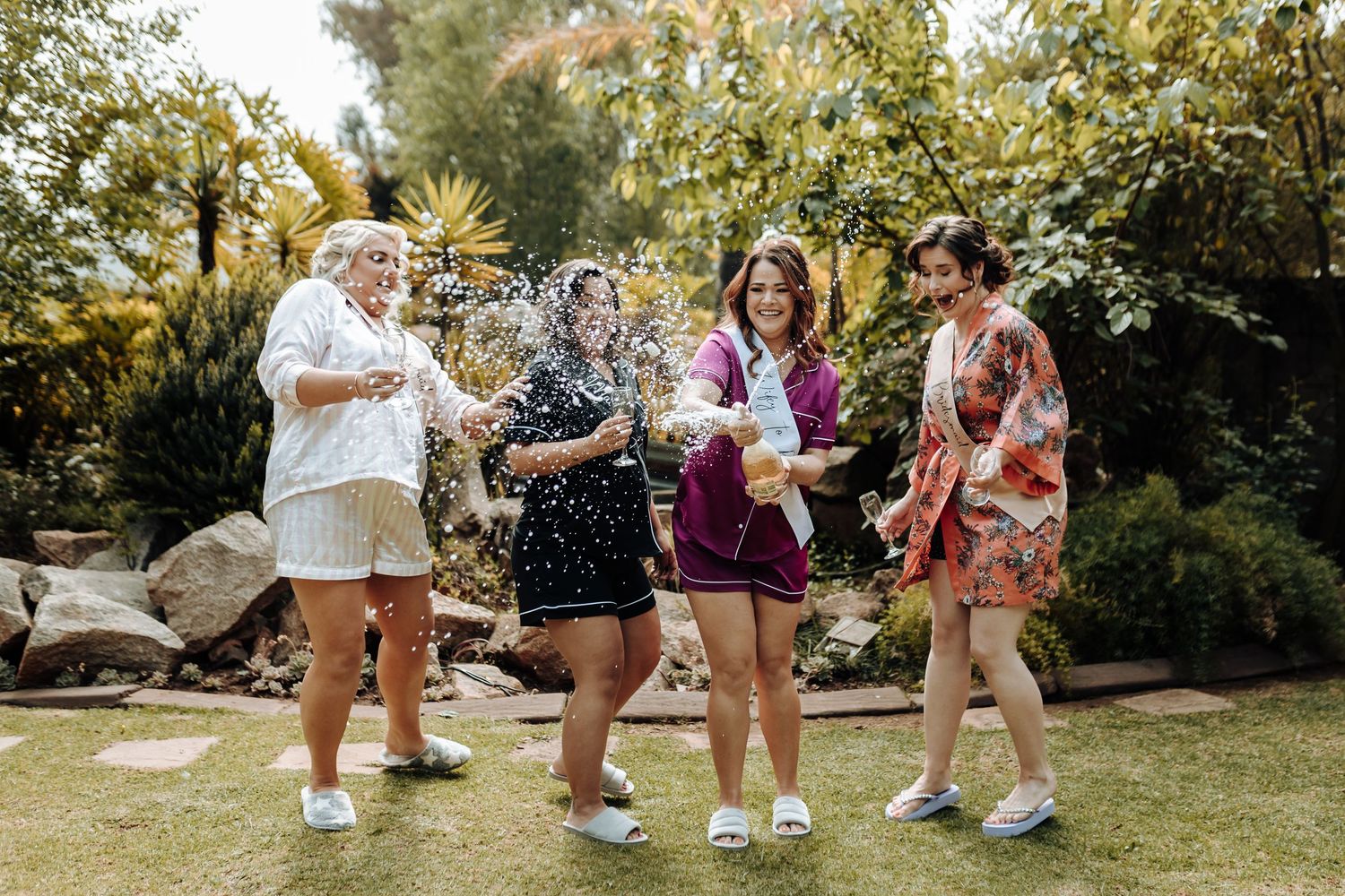Friends celebrate with champagne sprays in a sunny garden during pre-wedding festivities.