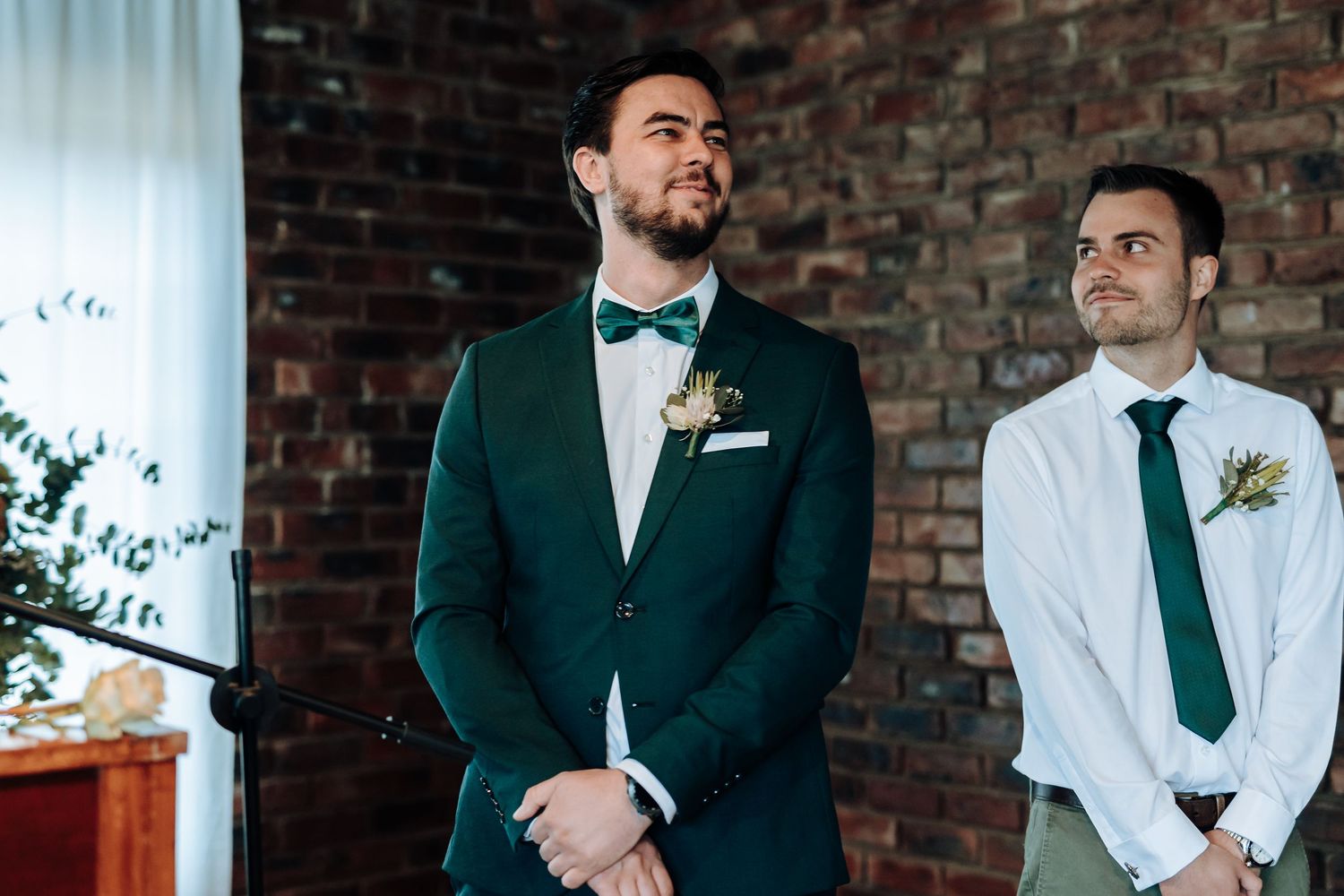 A groom in a forest green tuxedo with bow tie stands at the altar during a wedding ceremony.