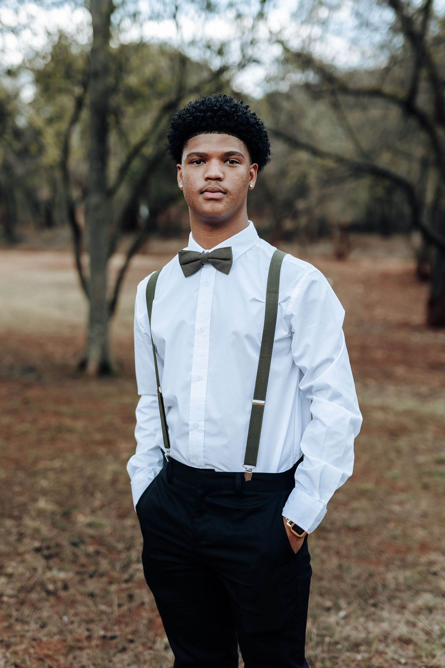 Young man in formal white dress shirt, black bow tie and suspenders poses in woodland setting.