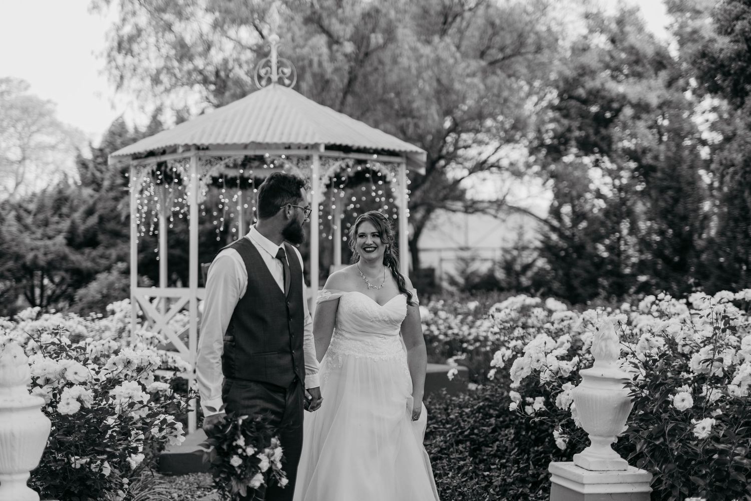 Black and white wedding photo of a couple standing by a gazebo in a rose garden.