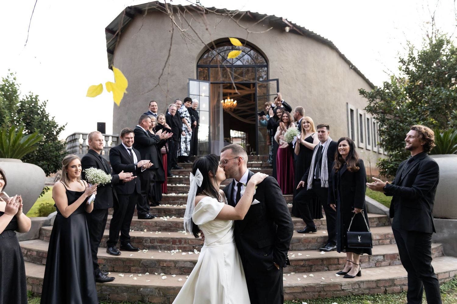 Wedding party celebrates with newlyweds on church steps during outdoor ceremony celebration.