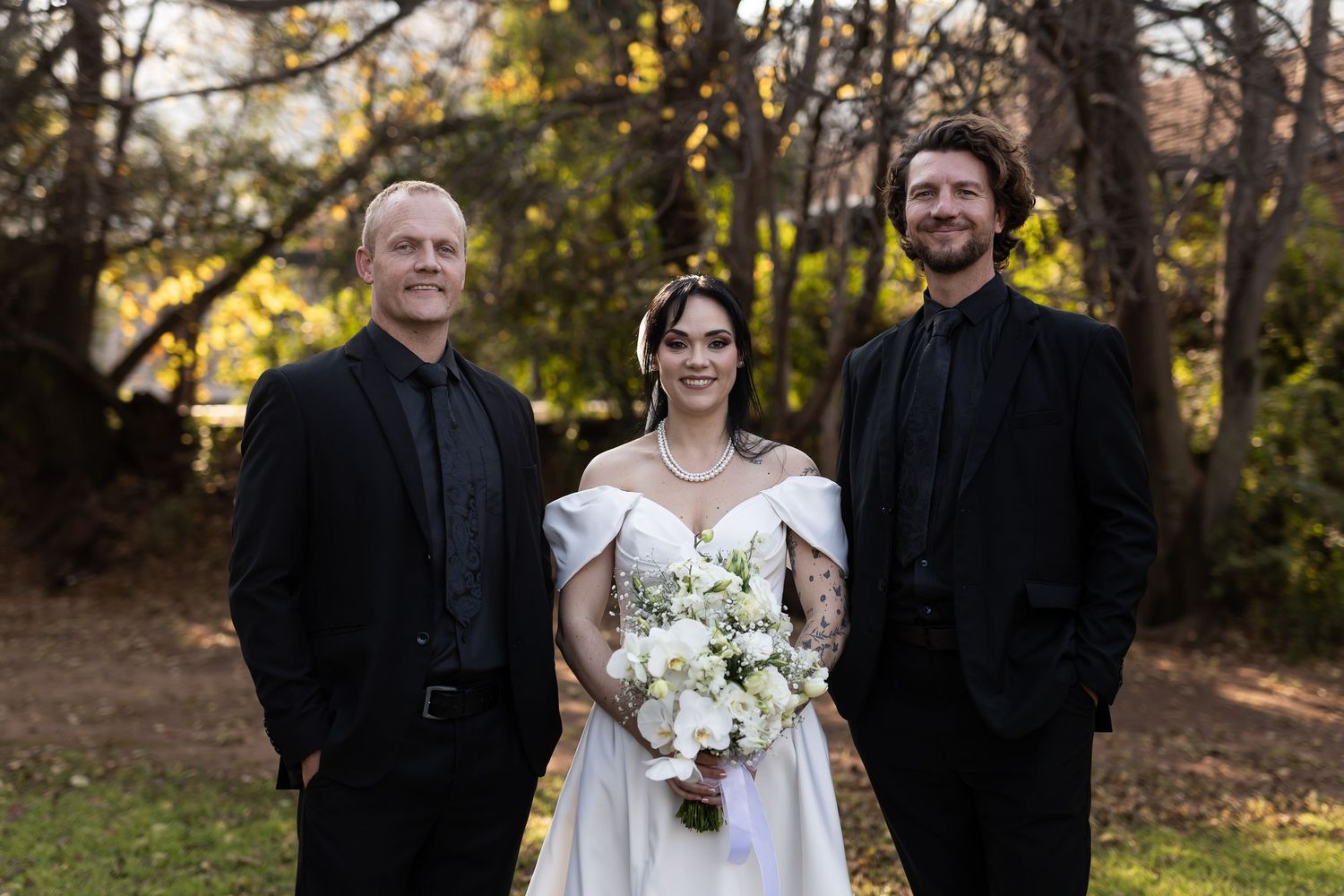 Outdoor wedding photo with bride in white dress holding bouquet standing between two people in black attire at sunset.