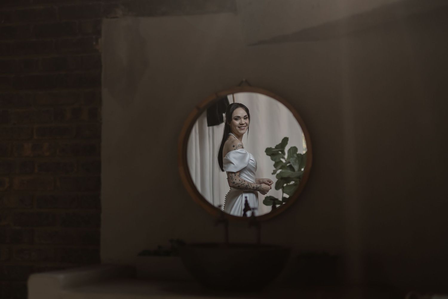 Moody reflection in circular mirror shows someone in white standing by houseplant in dark room.