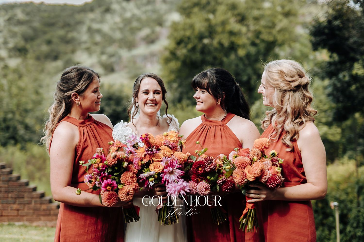 Bride with three bridesmaids in rust-orange dresses holding vibrant floral bouquets.