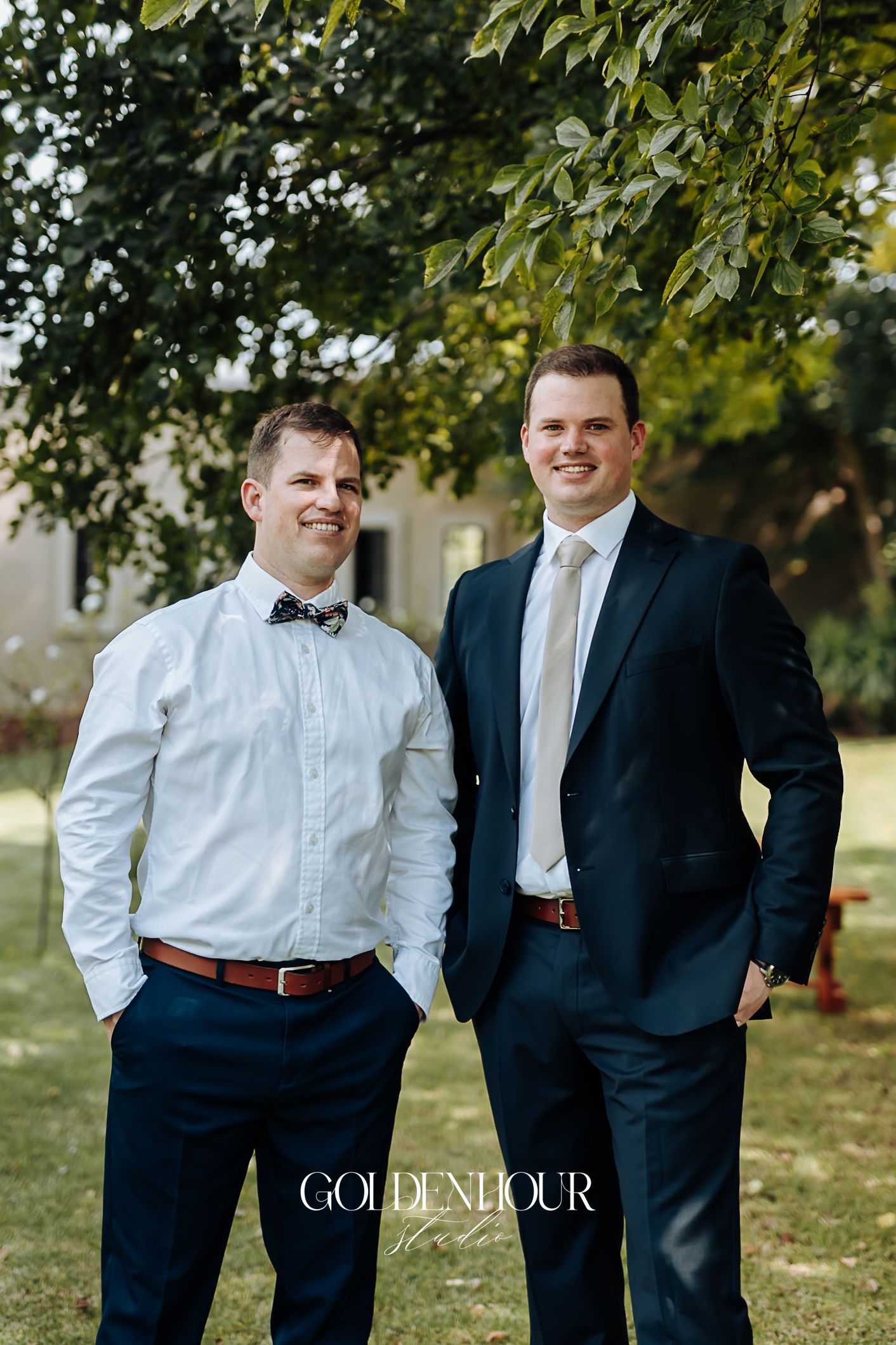 Two groomsmen in navy suits smiling outdoors under a leafy green tree at a wedding.