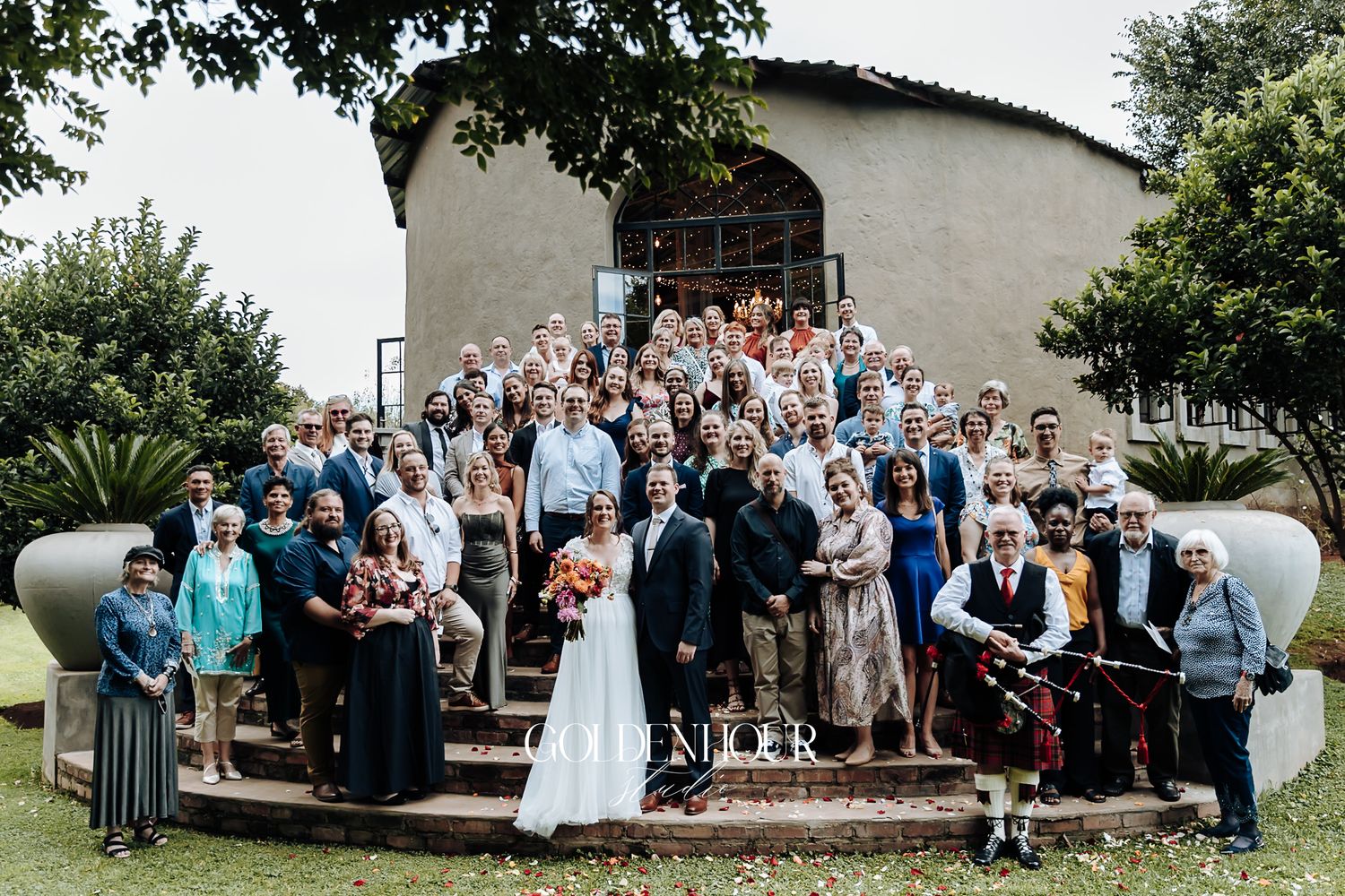 Large wedding group photo on stone steps outside a rustic venue with lush greenery.