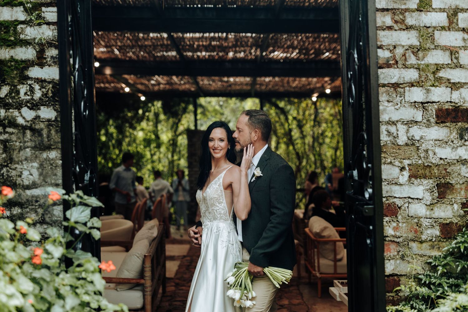Bride and groom pose together under a rustic brick archway surrounded by lush greenery at an outdoor wedding venue.