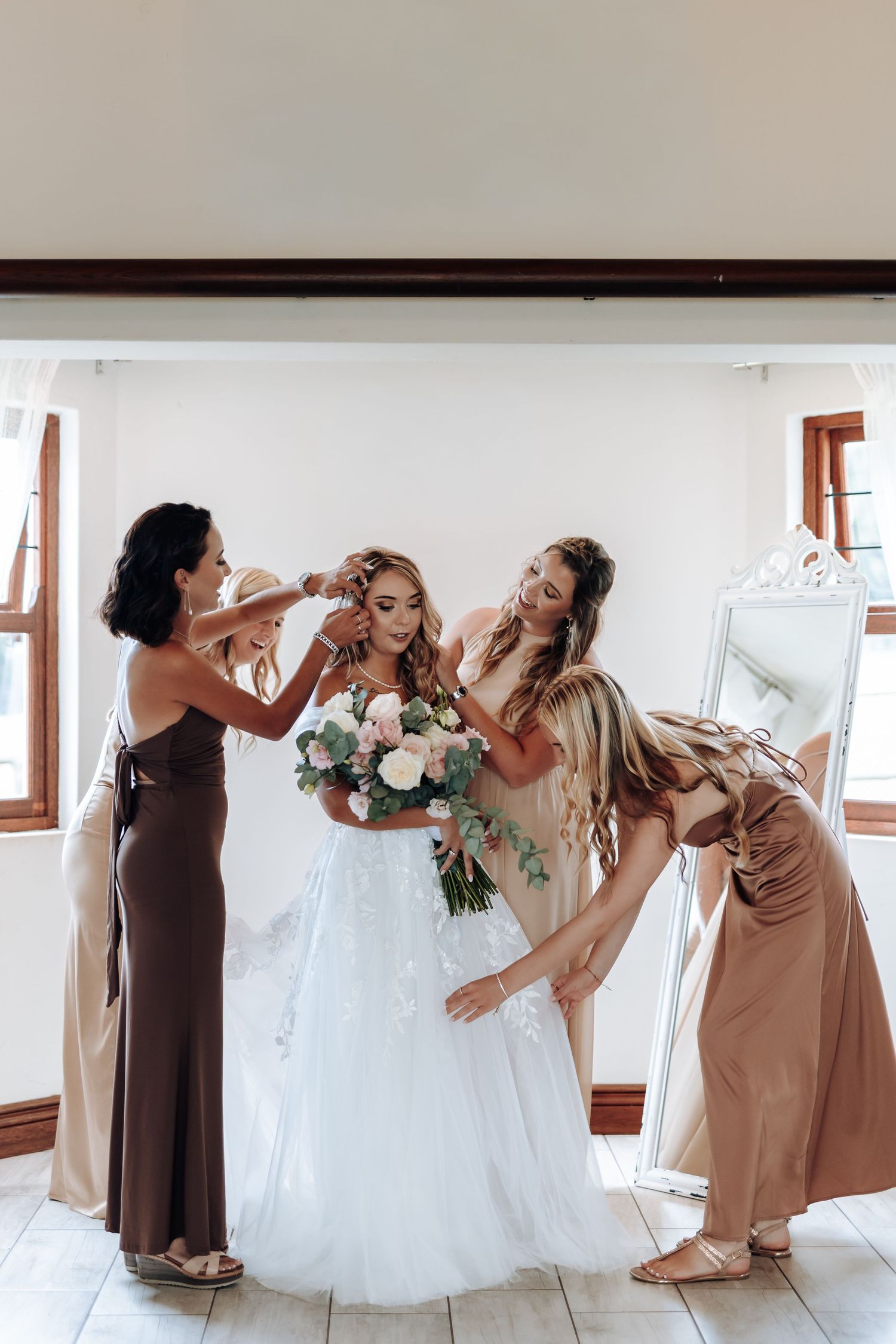 Bridesmaids in beige dresses help adjust the bride's white gown in a bright indoor setting during wedding preparations.