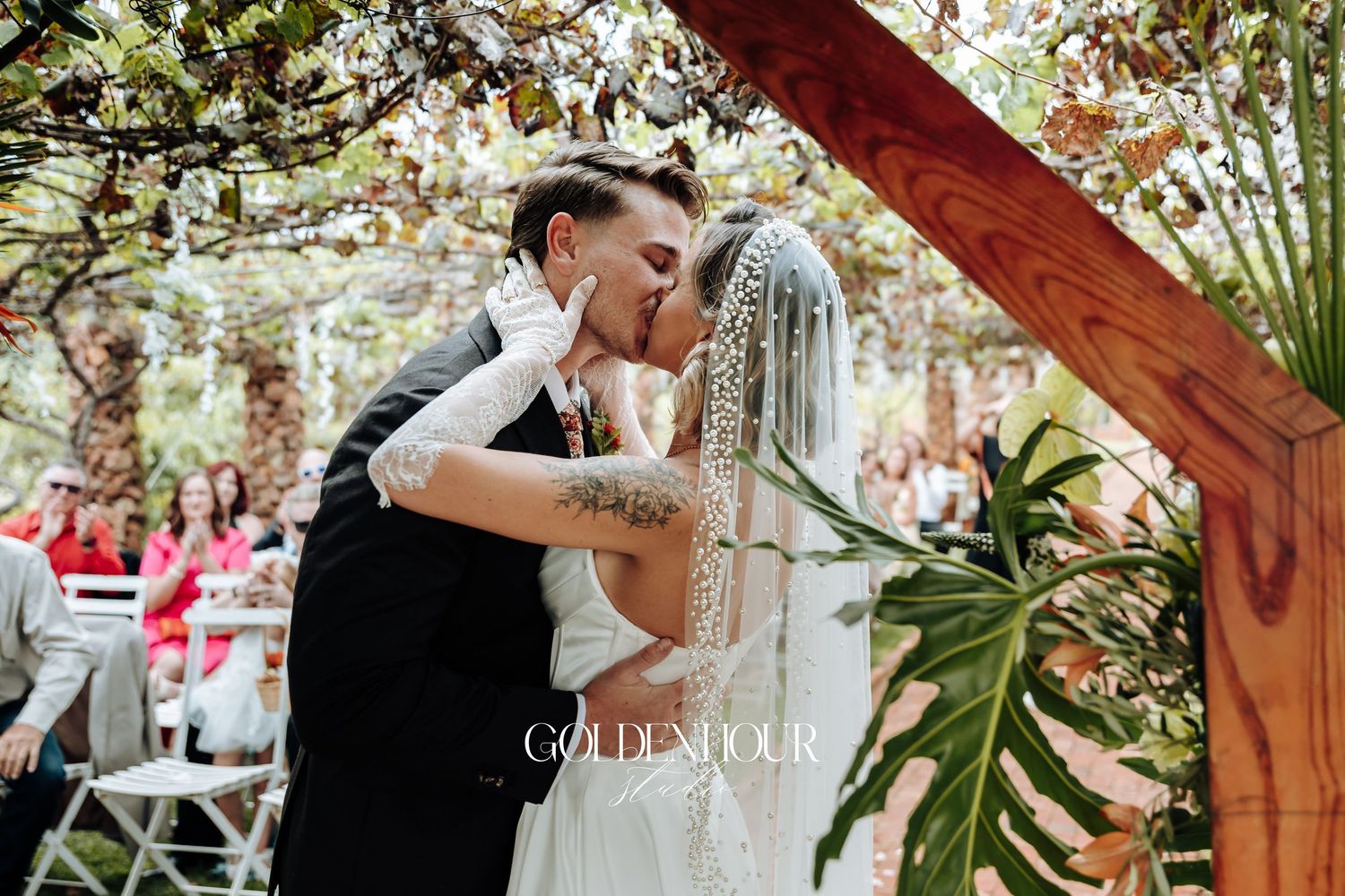 Wedding ceremony under wooden pergola decorated with tropical greenery.