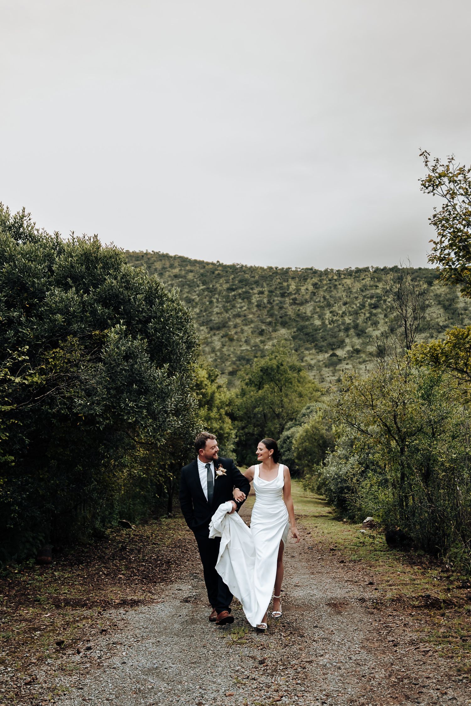 A couple walks hand in hand on a dirt path surrounded by green hills and trees in a romantic outdoor setting.