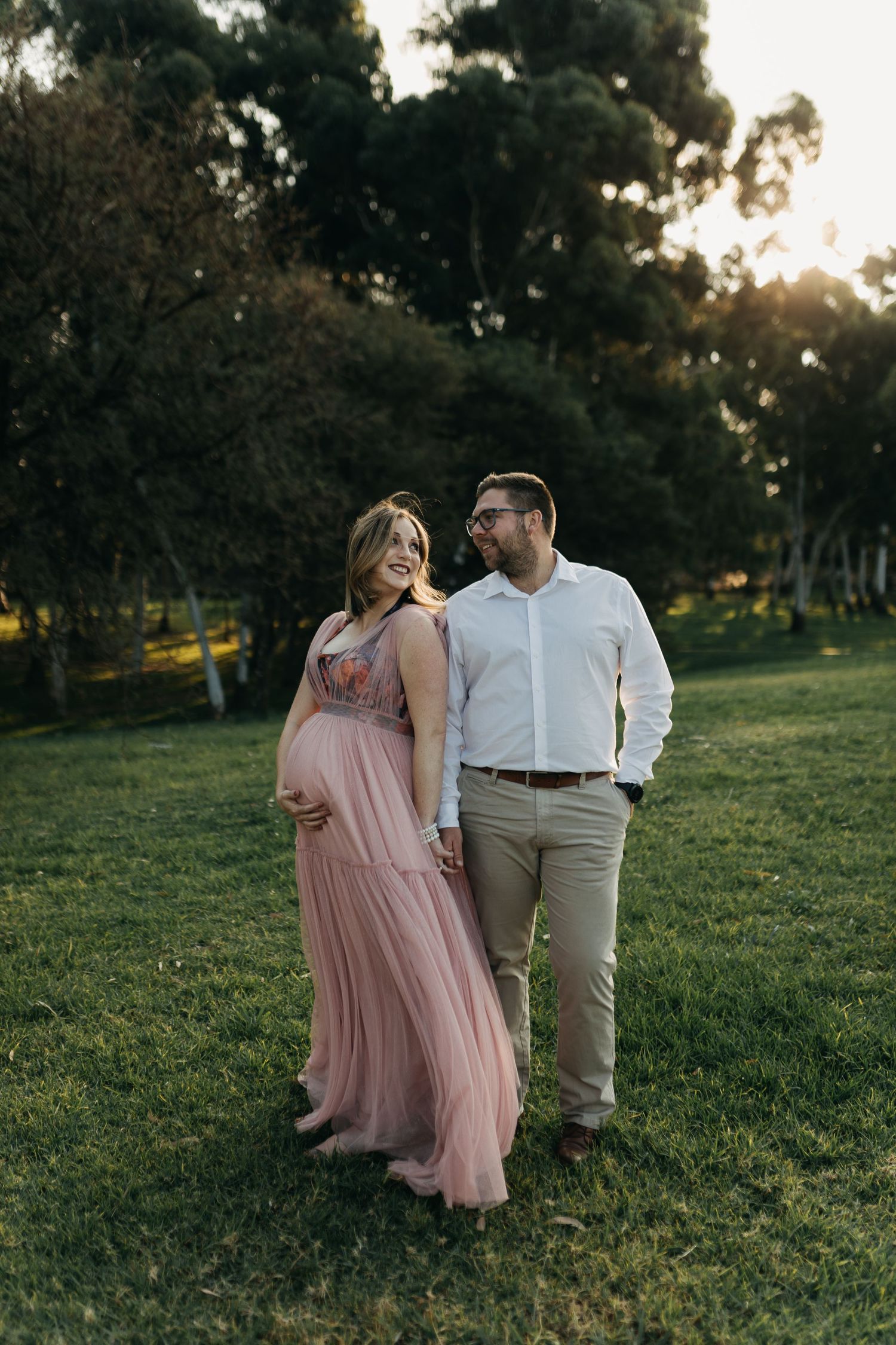 A romantic couple standing in a grassy field at sunset, with the person wearing a flowing pink dress and white shirt.