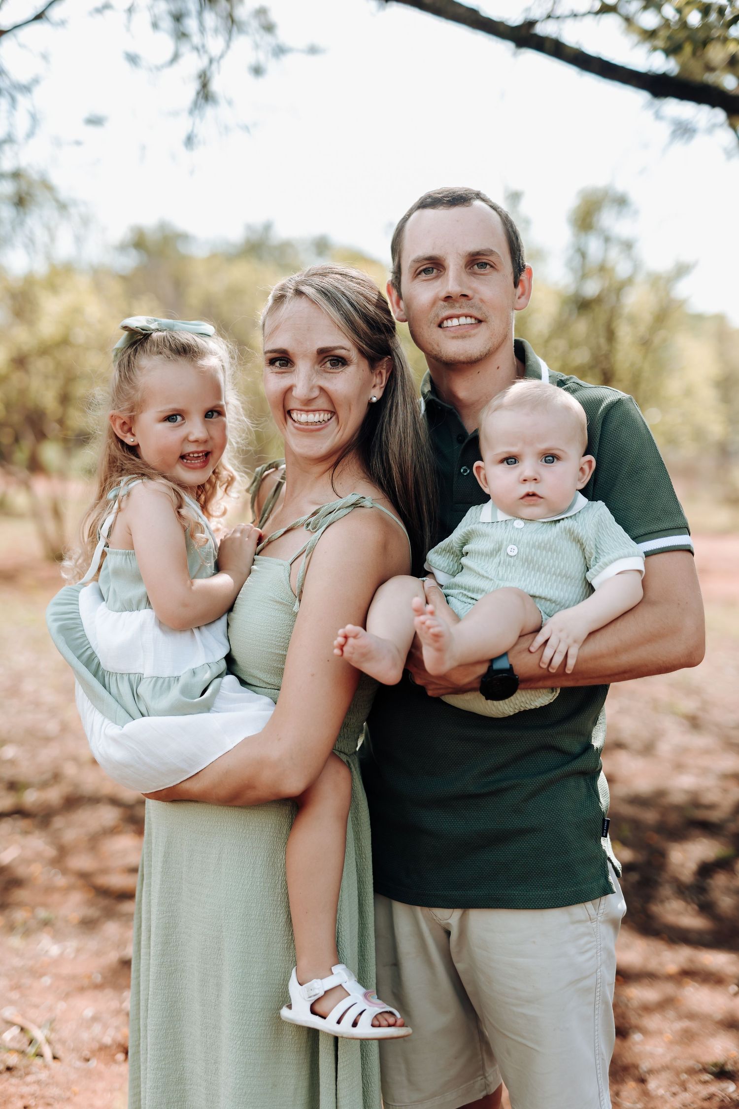 A young family in coordinating sage green and white outfits poses together in a natural outdoor setting.