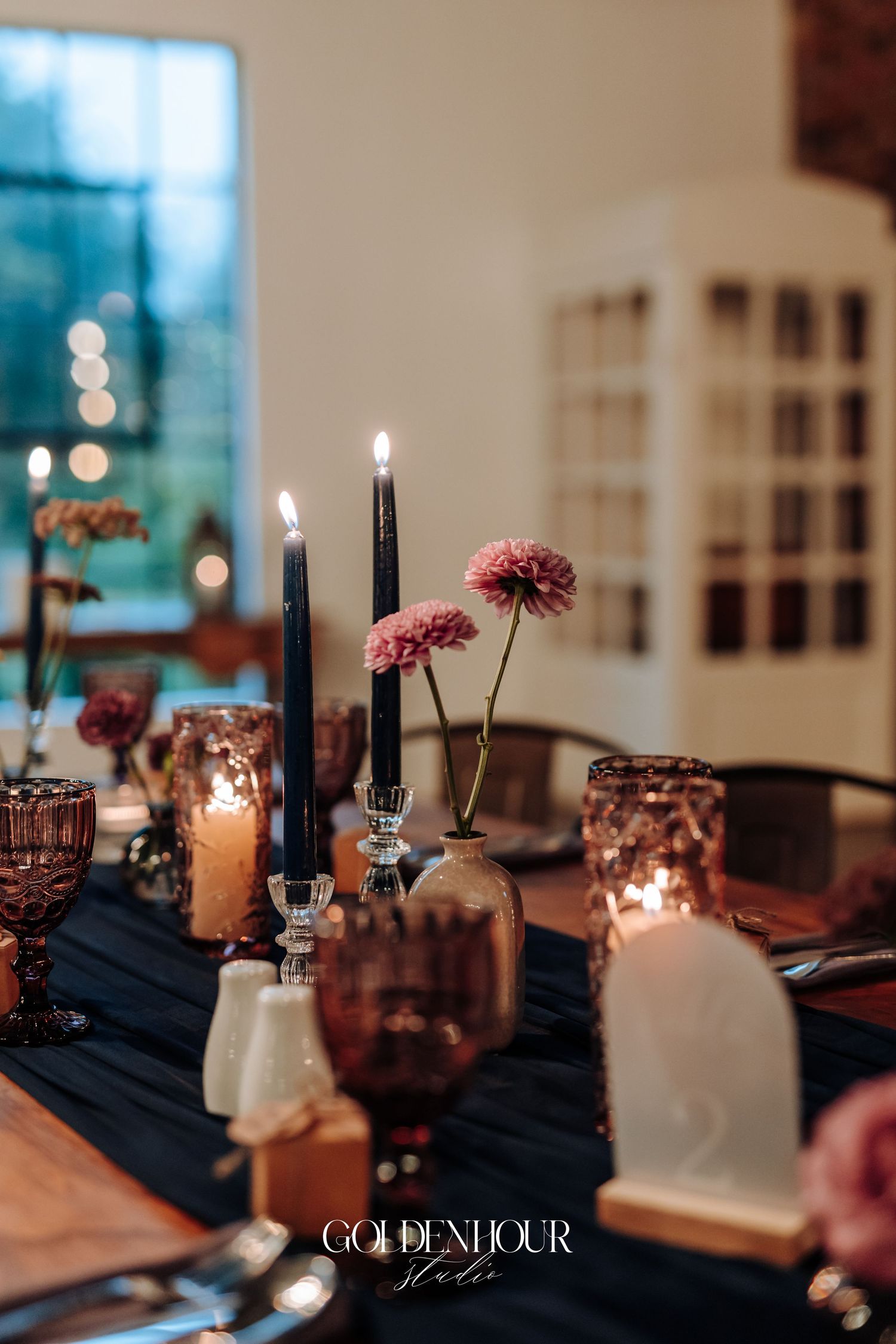 Elegant wedding table setting with dark candles, pink flowers, and moody purple glassware on navy runner.