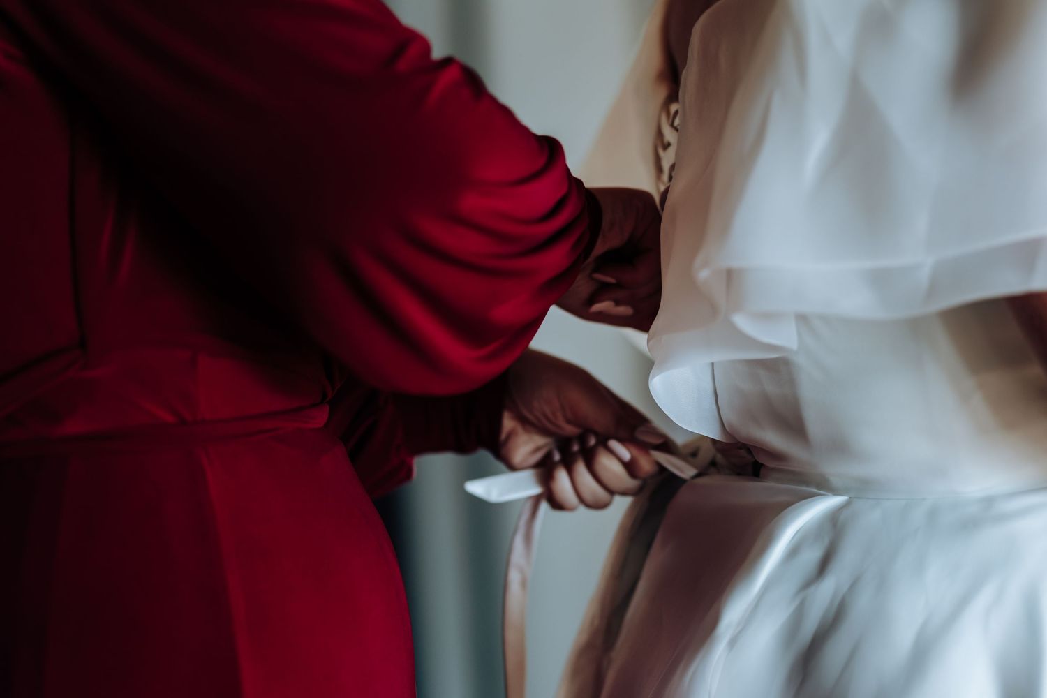 A bride in white gown being helped with her ribbon belt by a bridesmaid in a red dress.