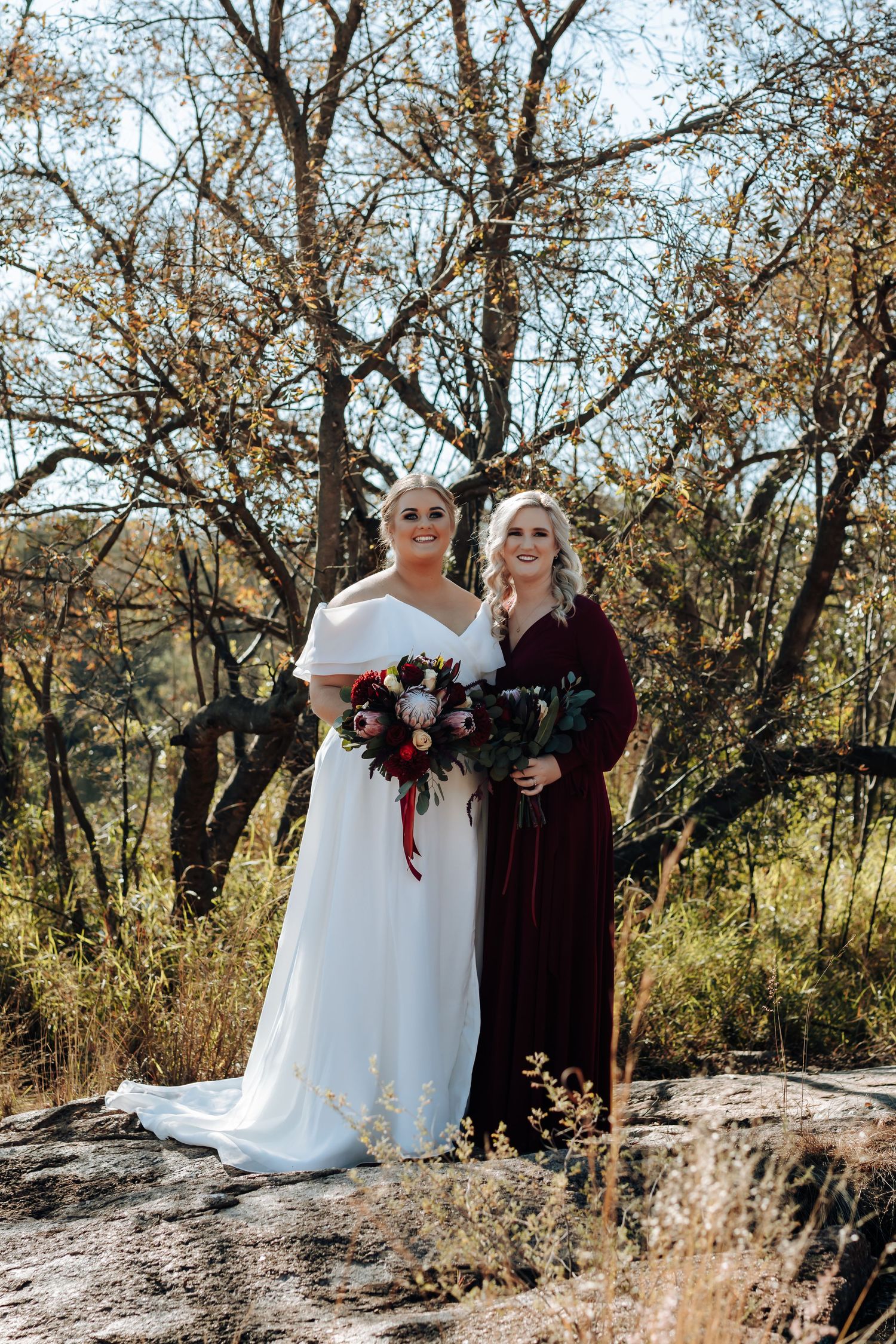 Bride in white off-shoulder gown and bridesmaid in burgundy dress holding bouquets outdoors among autumn trees.