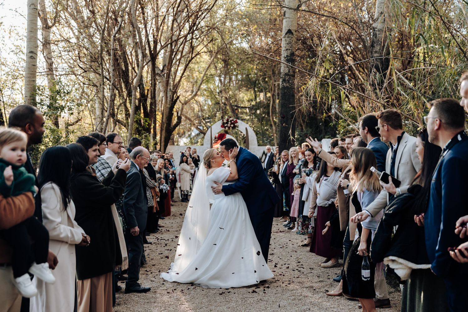 Bride and groom share a kiss while guests celebrate along a tree-lined aisle at an outdoor wedding ceremony.