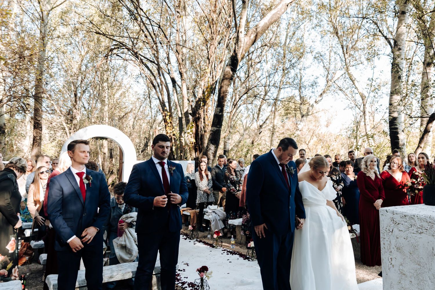 Outdoor wedding ceremony in a wooded setting with bride, groom, and officiant at the altar surrounded by guests.