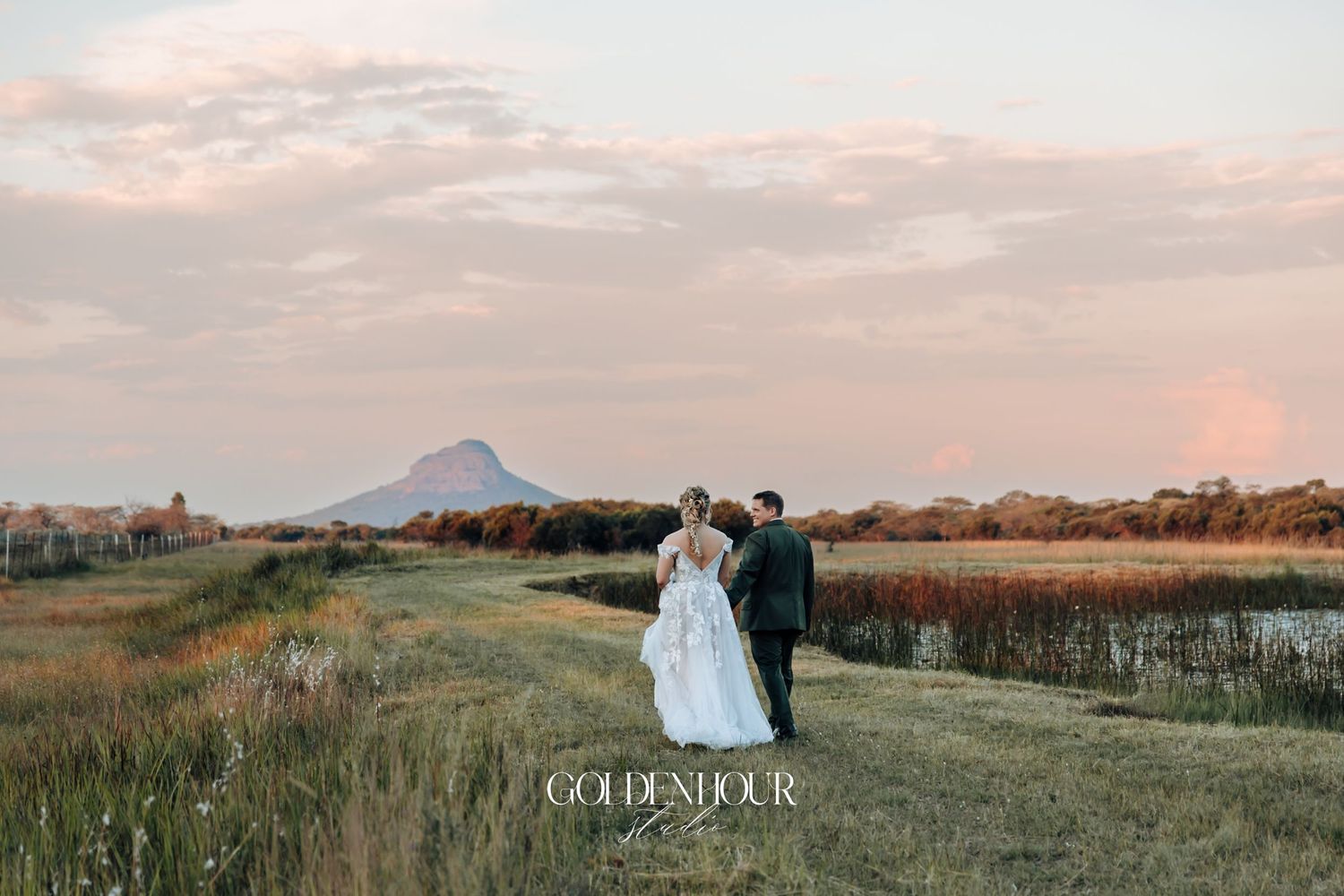 Bride and groom walking hand in hand along a golden field path at dusk with a distant mountain.