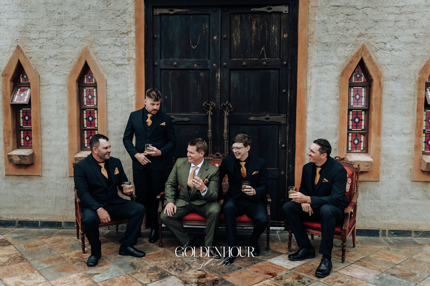 Five groomsmen in black and olive suits socializing outside ornate dark wooden doors at wedding venue.