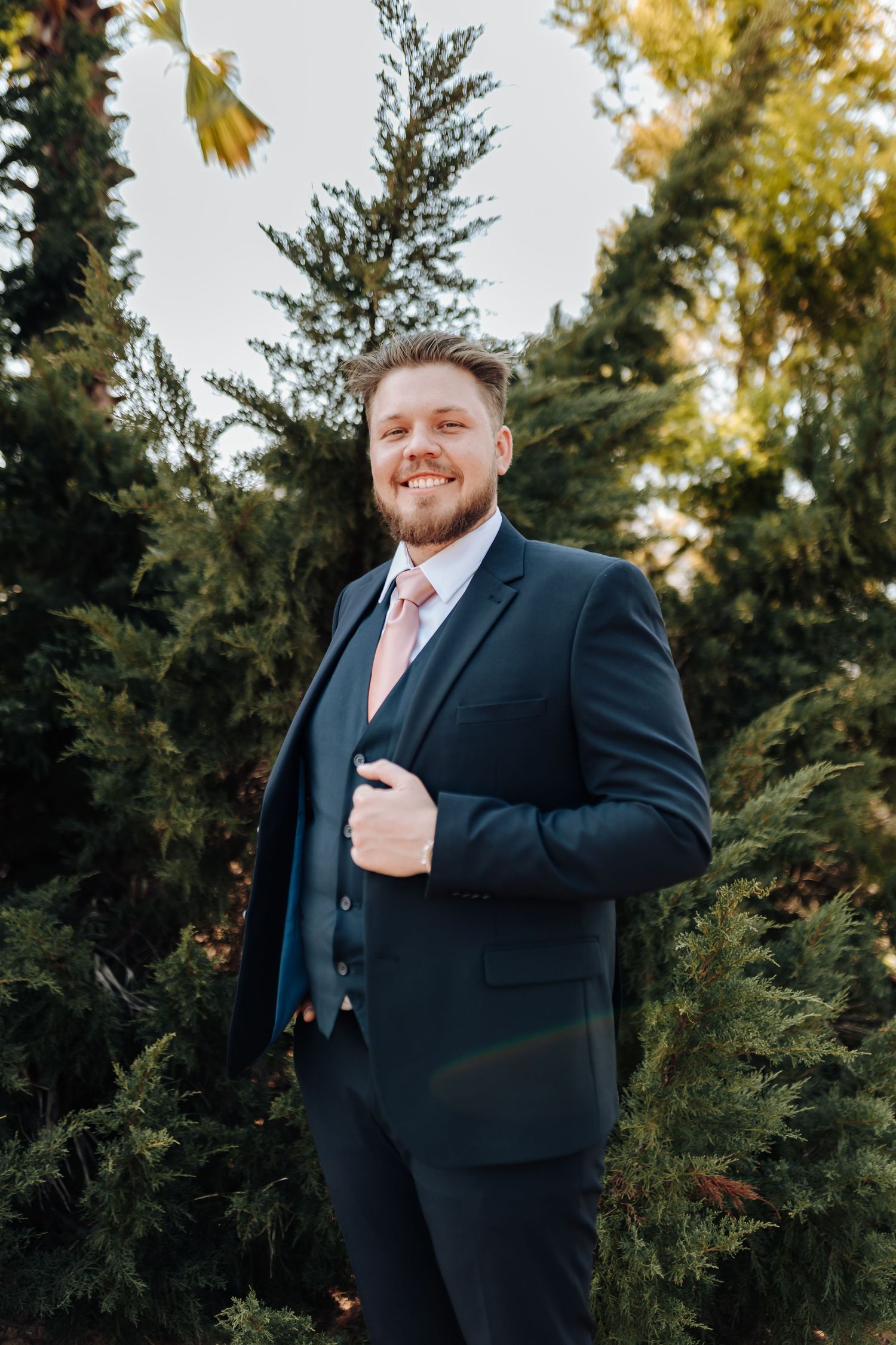 A person in a navy blue suit with a salmon pink tie poses confidently against an evergreen backdrop during golden hour.