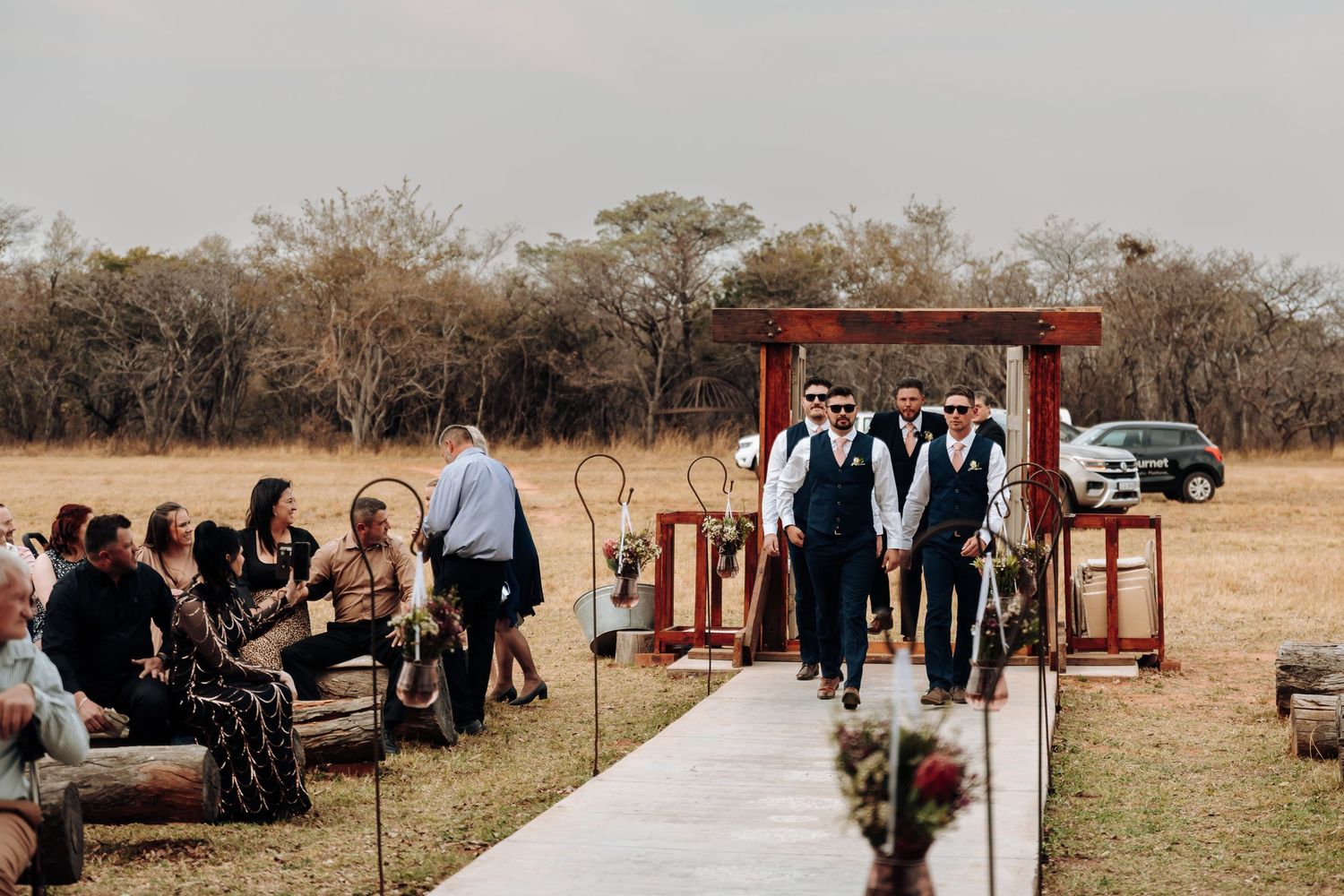 Outdoor wedding ceremony setup with rustic wooden arch and white aisle runner surrounded by guests in a rural setting.