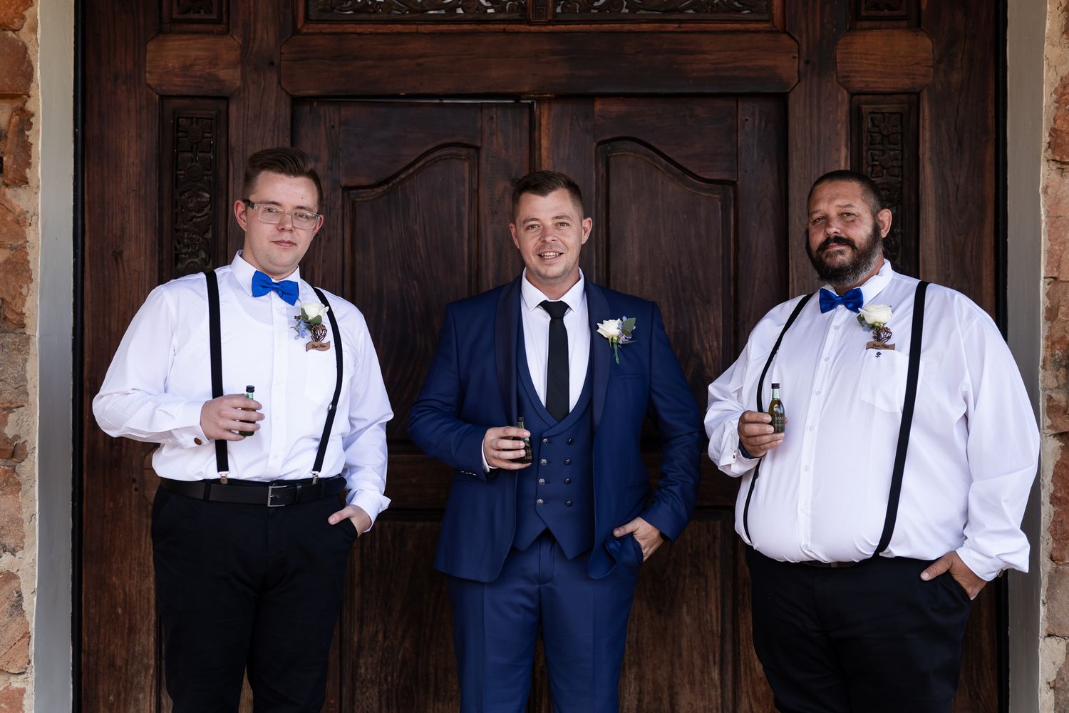 Three groomsmen in formal attire pose together in front of wooden doors.