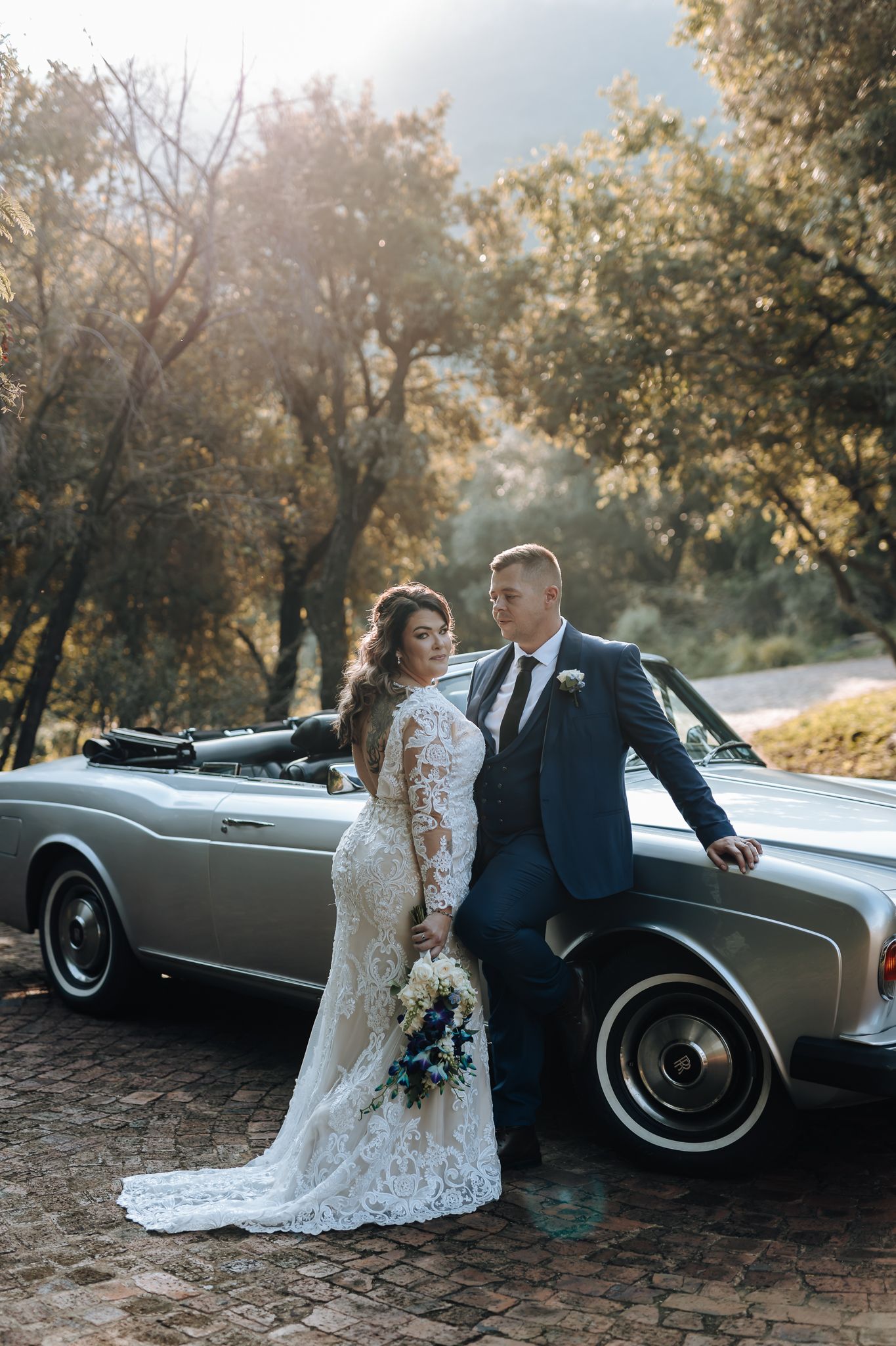 Bride in lace gown and groom in suit pose with vintage car during golden hour.