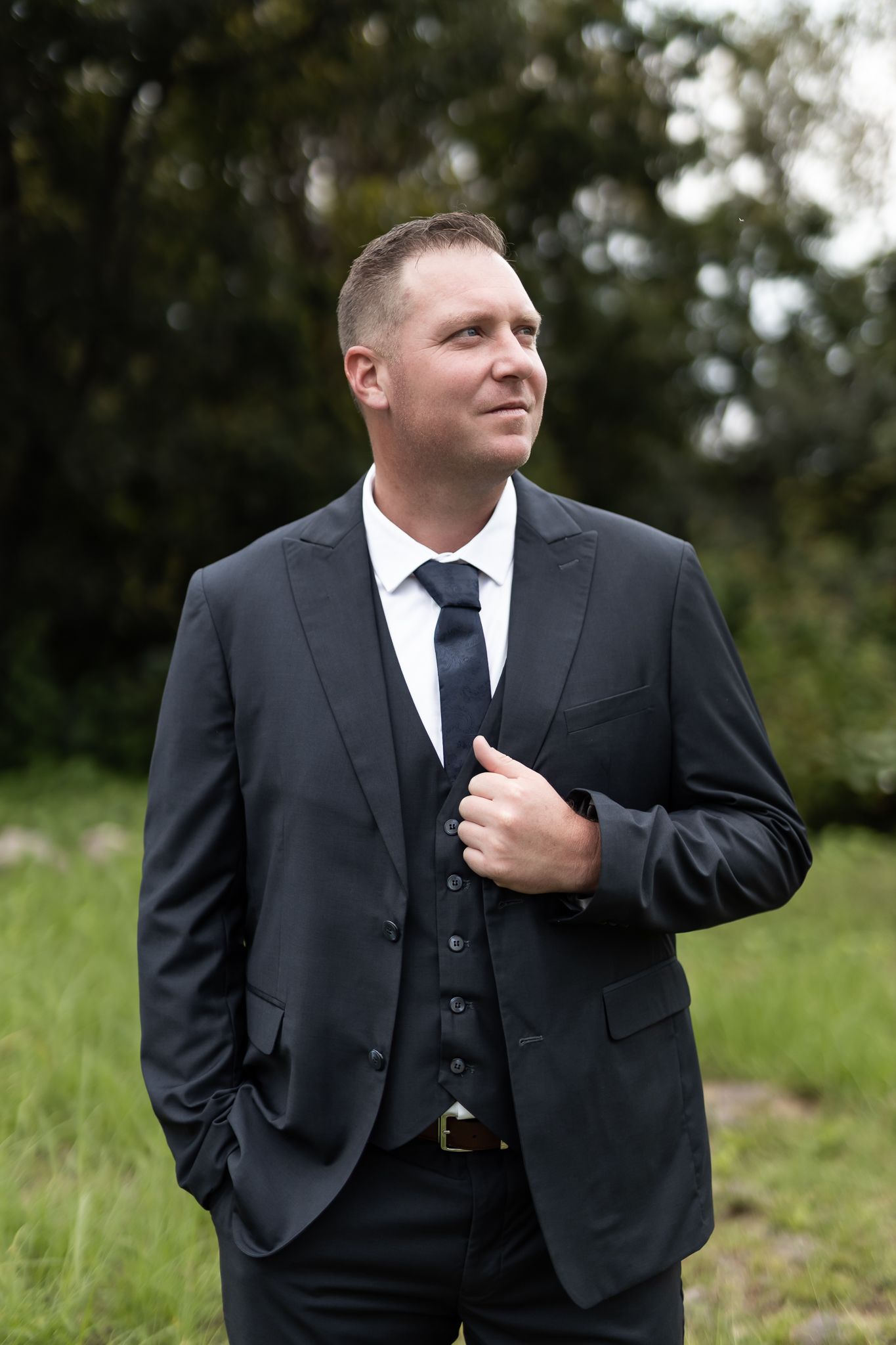A groom in a classic black three-piece suit adjusts his jacket in an outdoor setting.