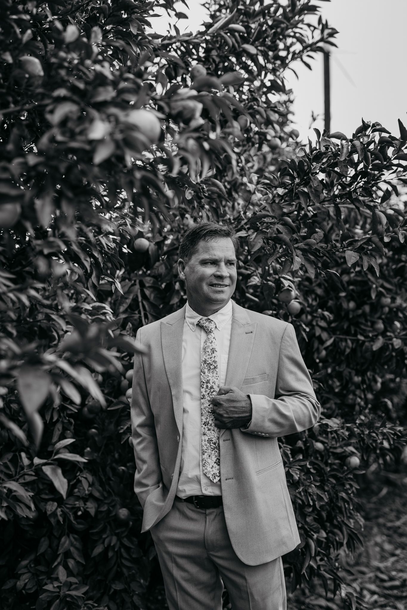 Wedding attendee in gray suit poses in black and white photo among orchard trees.