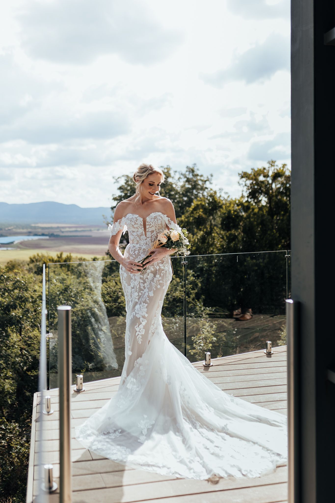 Bride wearing fitted lace gown with train stands on wooden deck overlooking mountain vista.