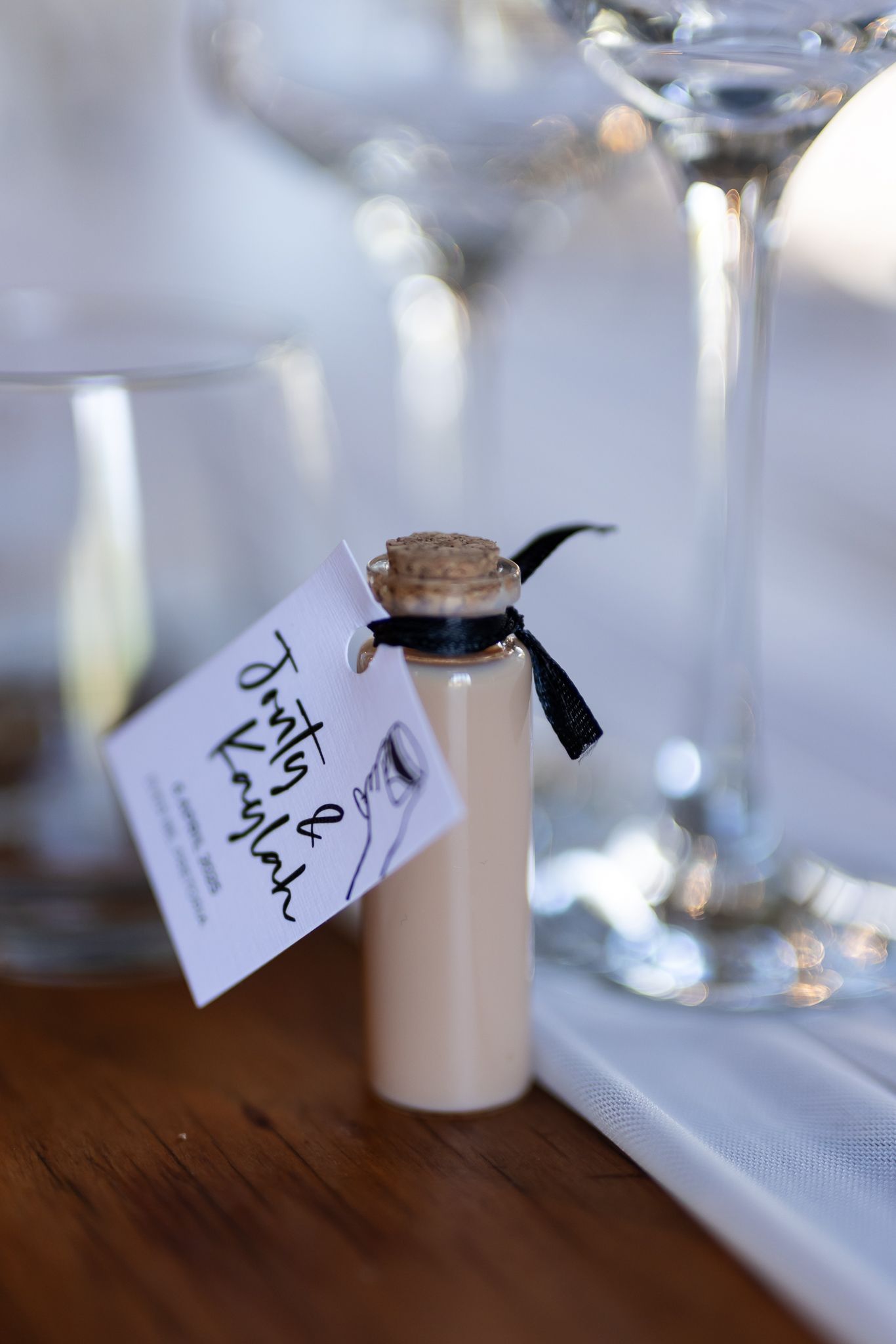 A small corked bottle with a black ribbon and tag sitting beside wine glasses on a wooden table.