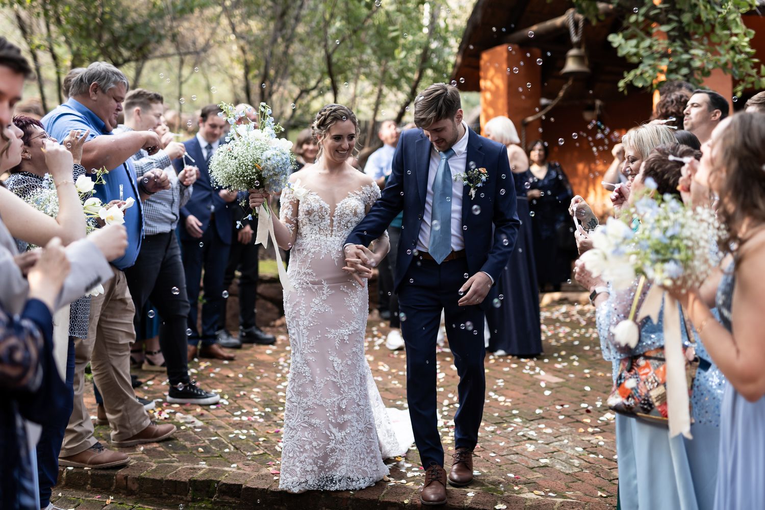 Wedding couple walks through guest-lined pathway while being showered with confetti in outdoor ceremony.
