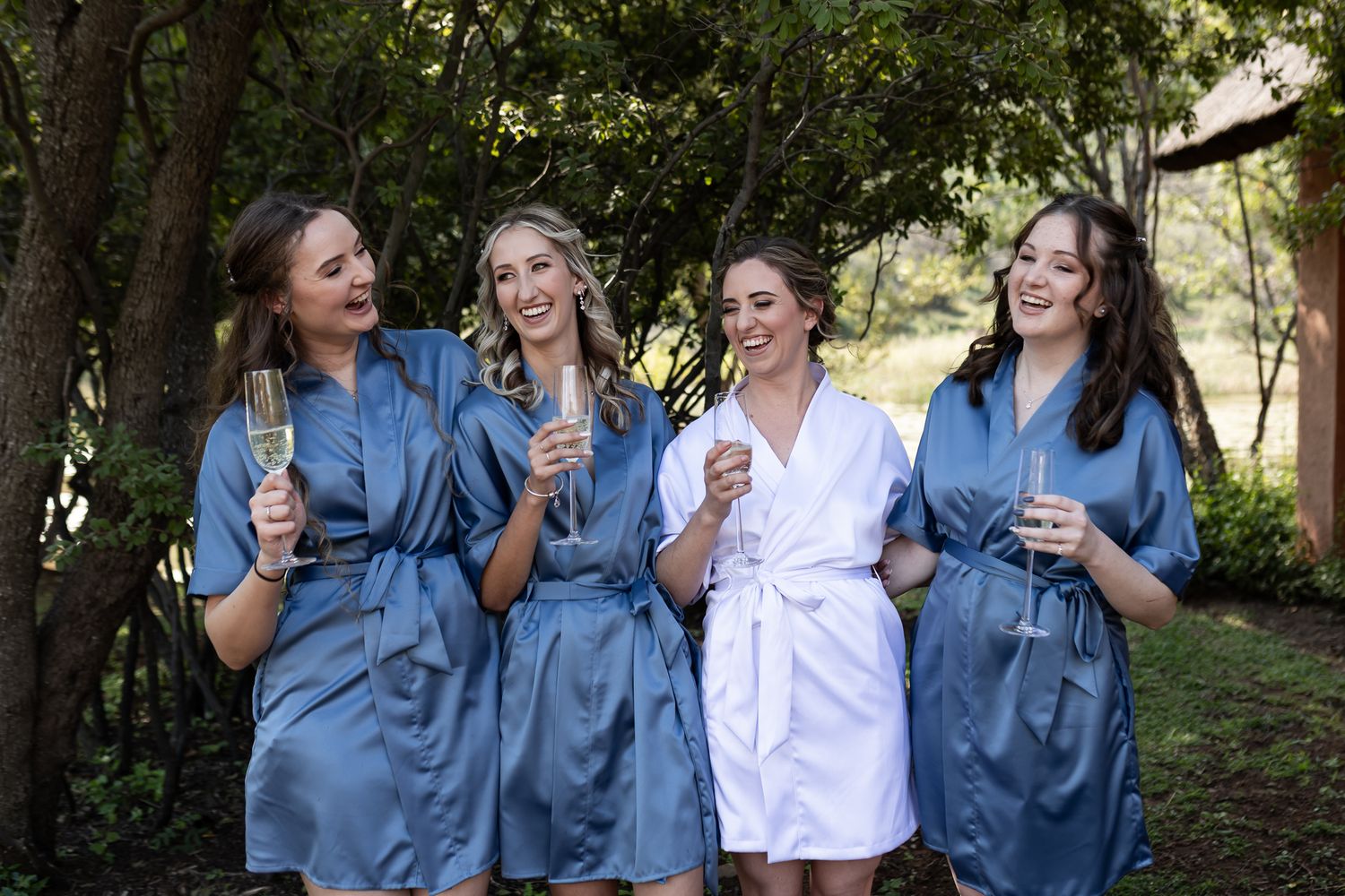 Friends in coordinating blue robes laugh while holding champagne glasses outdoors.