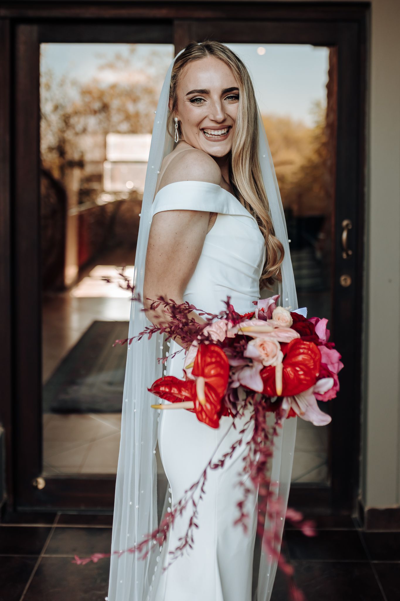 Candid bridal portrait with stunning red and pink cascading bouquet against glass doors.