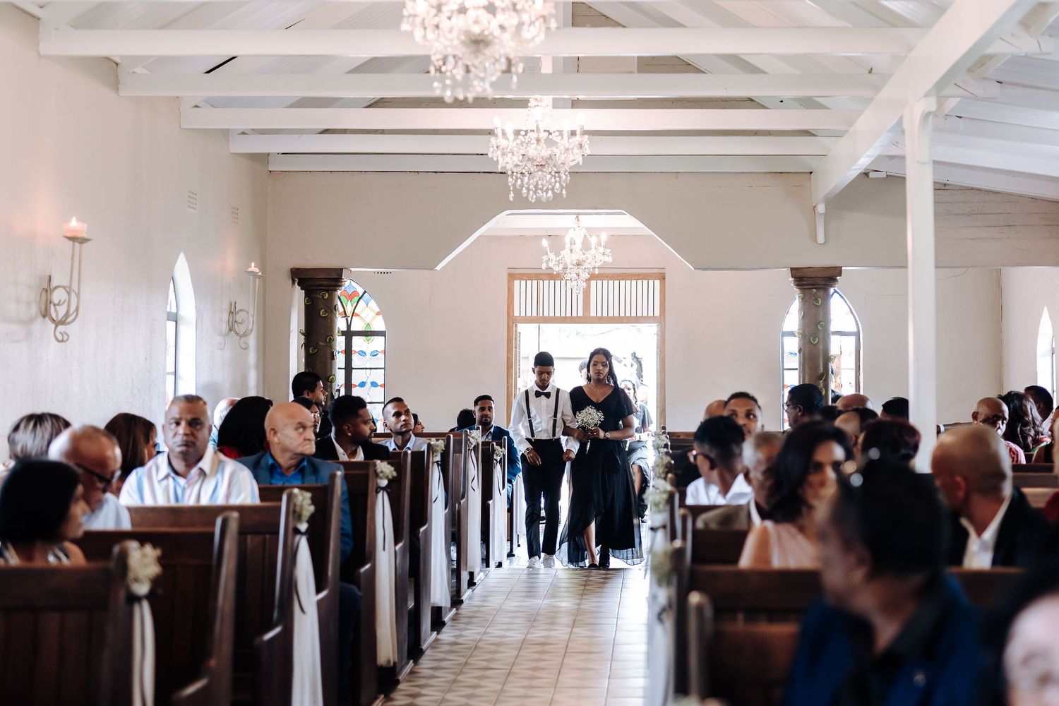 Wedding ceremony in progress inside elegant chapel with chandeliers and white beams.