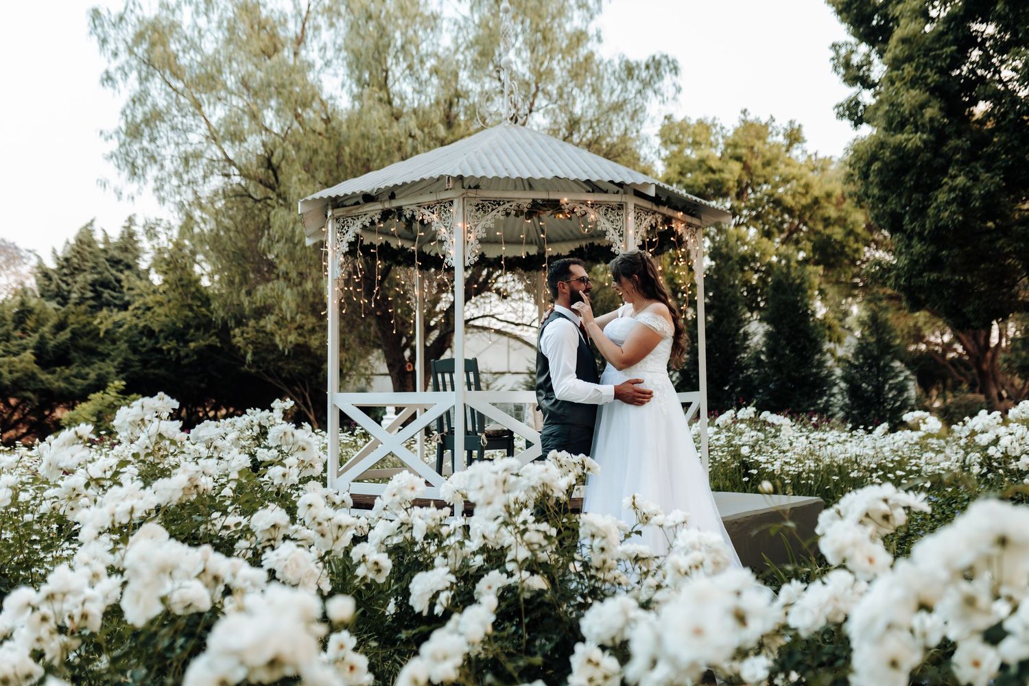 A romantic wedding moment in a white gazebo surrounded by blooming white flowers in a garden setting.