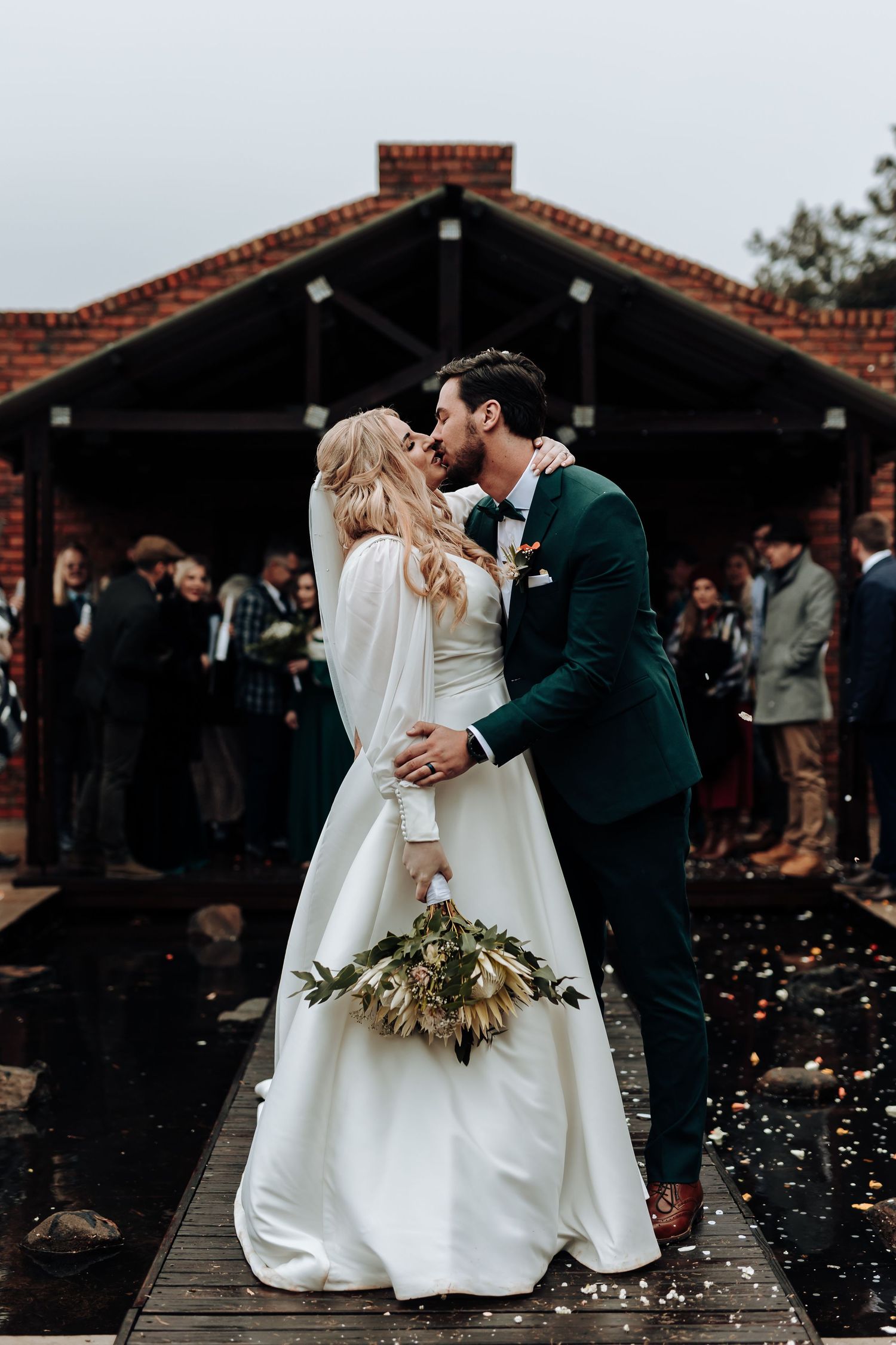 A romantic wedding kiss in front of a brick building with a dried floral bouquet.