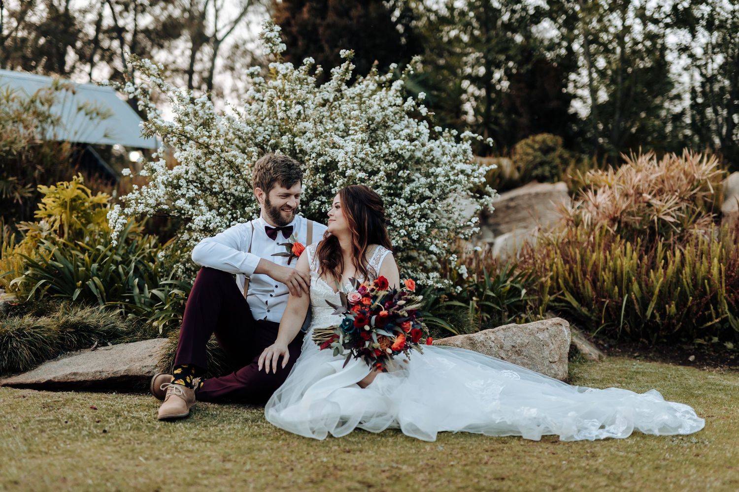 Intimate wedding portrait of couple sitting on grass with vibrant florals in background.