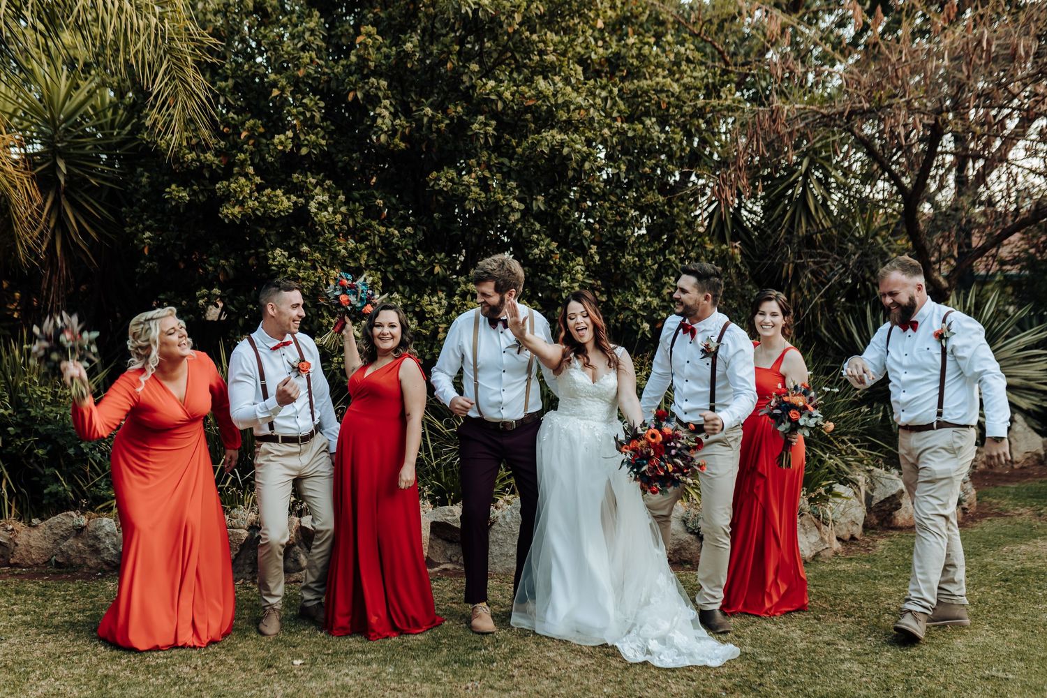 Casual outdoor wedding party portrait with bridesmaids in coral dresses and groomsmen in suspenders.