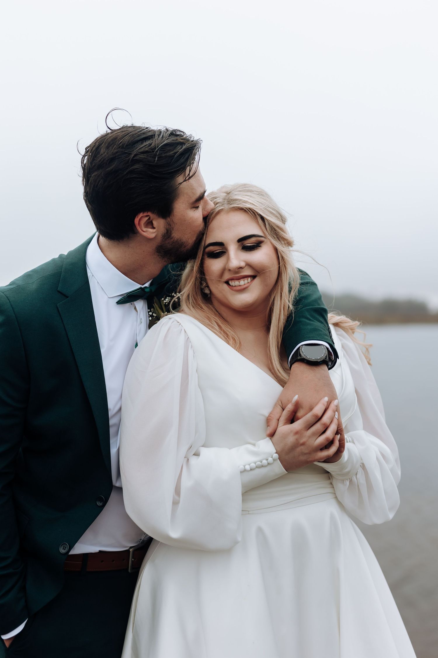 A tender moment between newlyweds in elegant wedding attire on a lakeside dock.